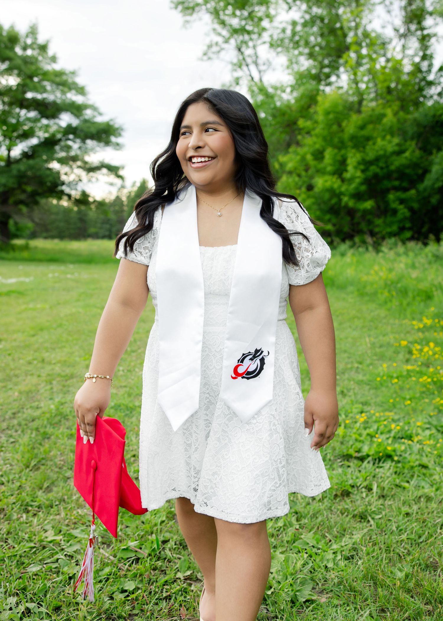 Smiling graduate in white lace dress and stole holds red cap outdoors in green field.