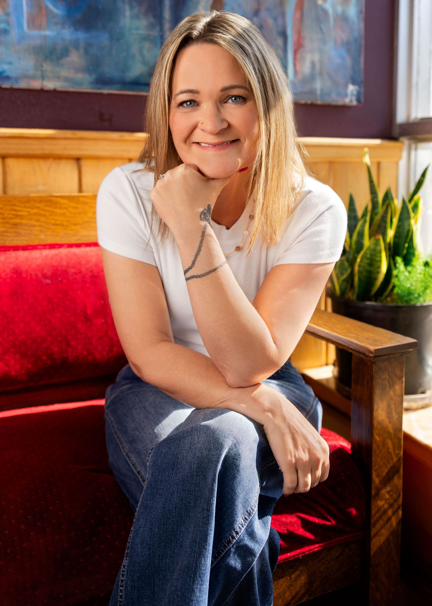 Smiling blonde woman in white t-shirt and jeans sitting relaxed in a cozy restaurant booth with plants.