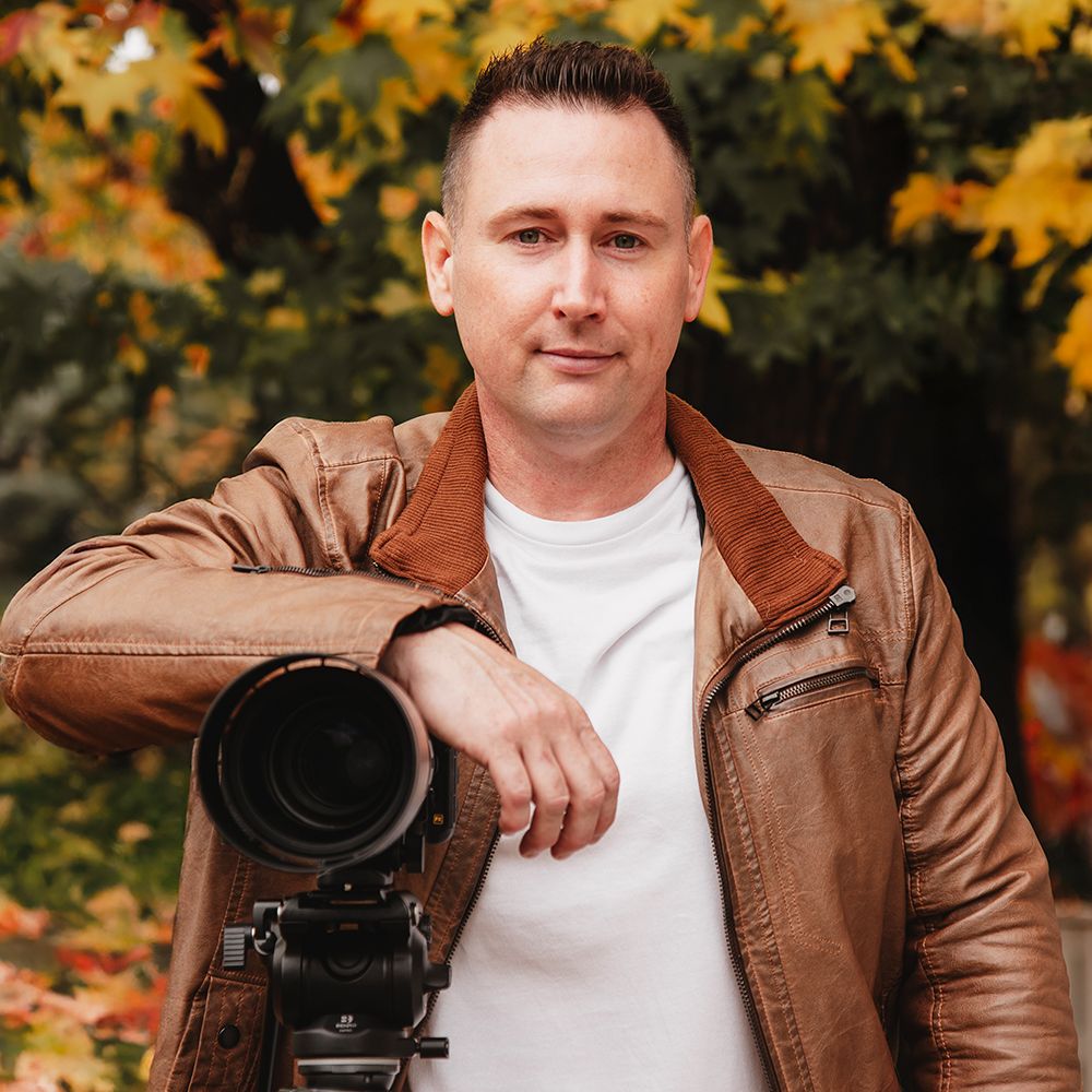 Man in brown leather jacket holding professional camera outdoors with autumn foliage background.
