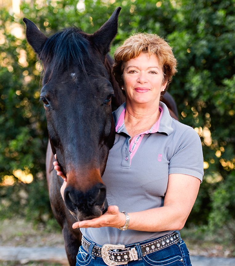 Woman in gray polo shirt poses with a black horse outdoors, smiling warmly while gently holding the horses muzzle.