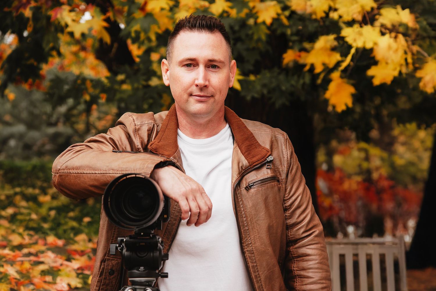 Man in brown leather jacket posing with camera on tripod among vibrant autumn foliage outdoors.