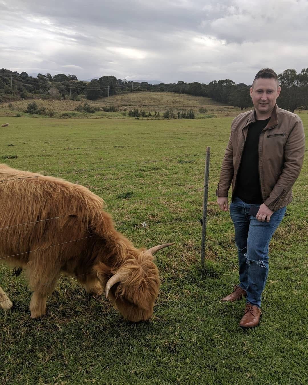 Man in brown jacket standing beside a fluffy Highland cow grazing in a green pastoral field under cloudy skies.