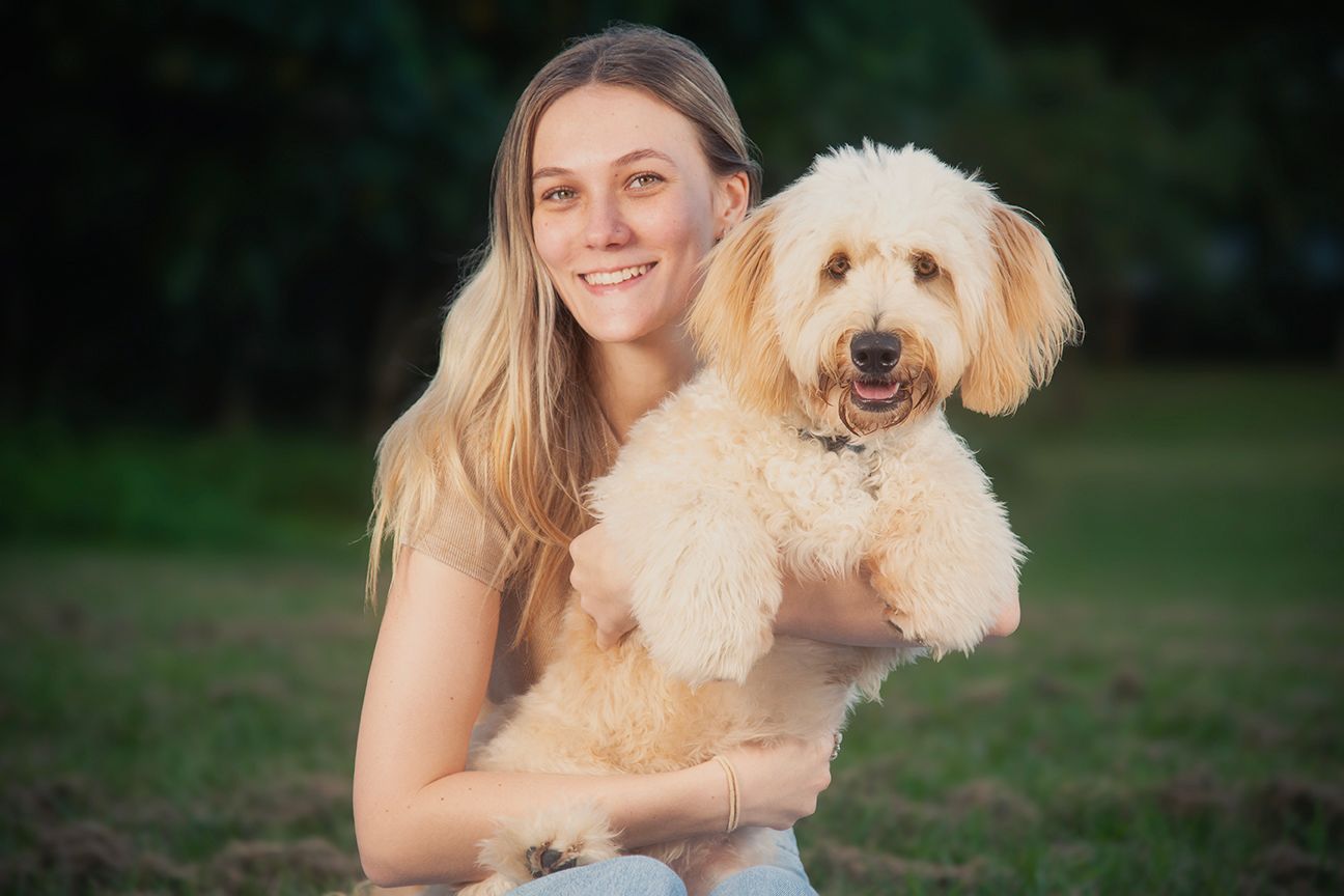 A person sits outdoors with a happy cream-colored Goldendoodle dog in a natural setting.