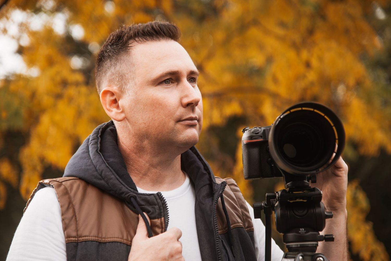 Photographer wearing brown vest holds professional camera against autumn foliage background.