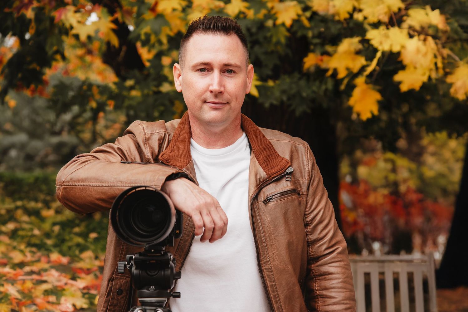 Professional photographer in brown leather jacket poses with camera equipment against autumn foliage background.