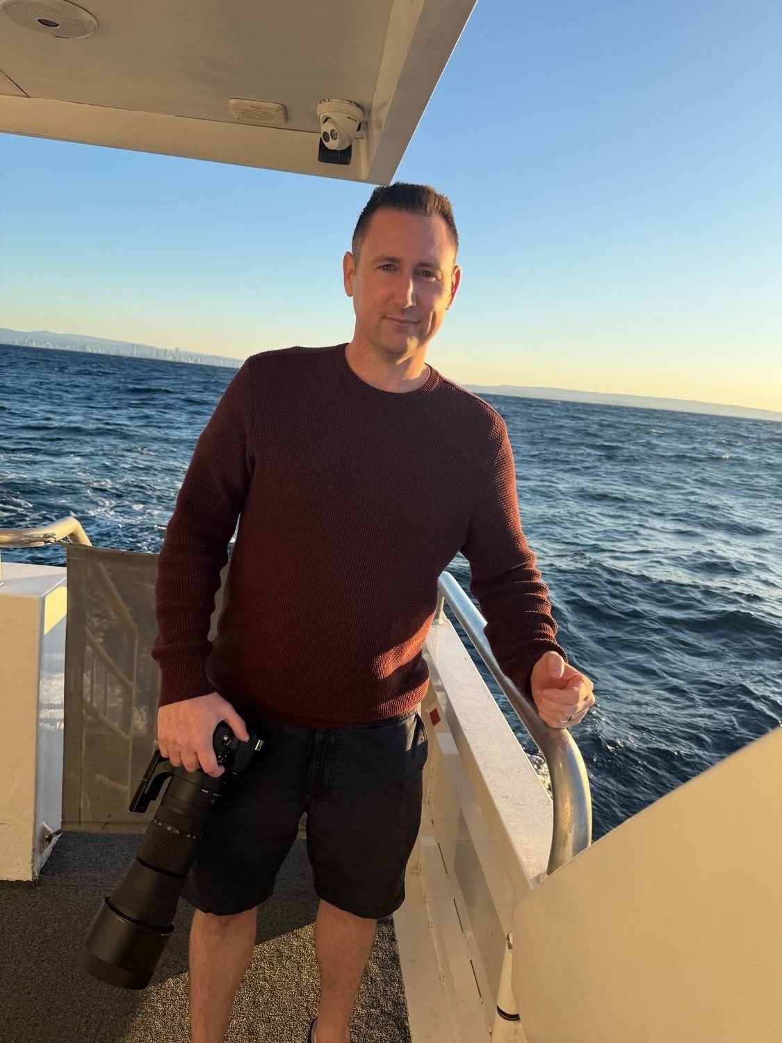 Man holding camera on boat deck with ocean view at sunset, wearing maroon sweater and black shorts.