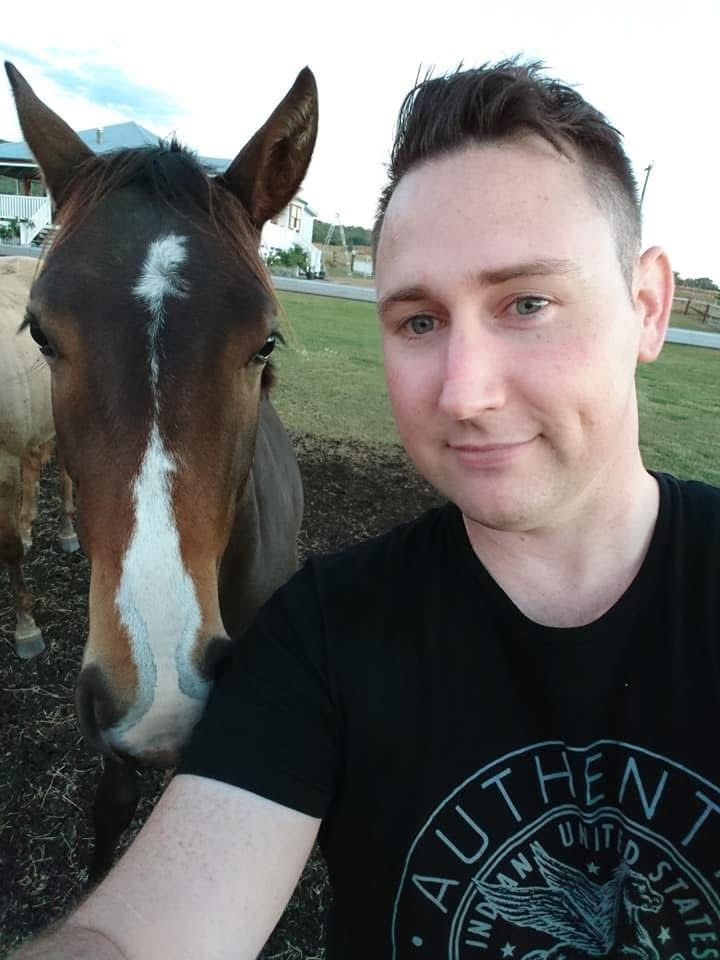 Young man taking a selfie with a brown and white horse outdoors near a farm, wearing an Authentic United States t-shirt.