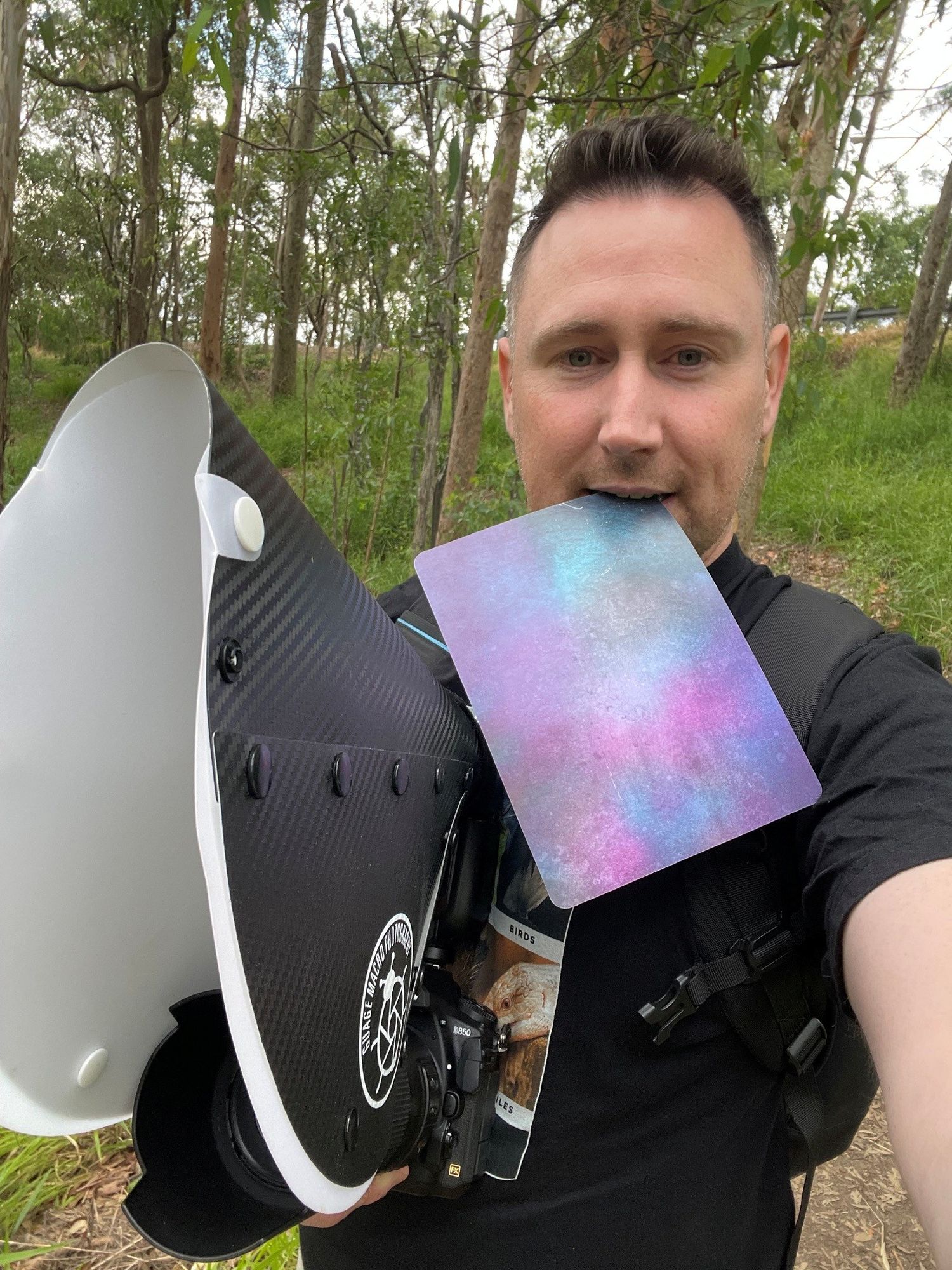 Man taking selfie outdoors holding carbon fiber wakeboard with colorful purple-pink fin, trees in background.