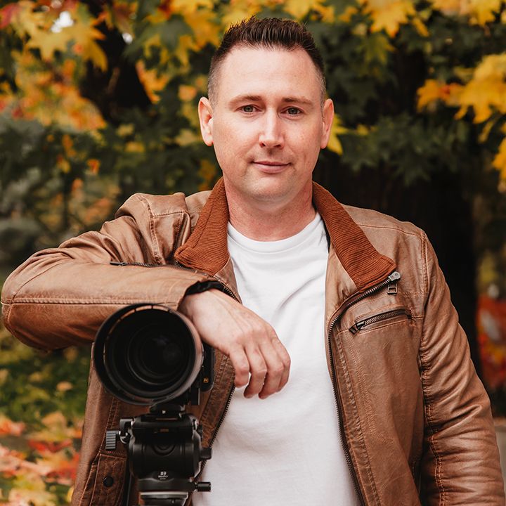Man in brown leather jacket leaning on professional camera with autumn foliage background.