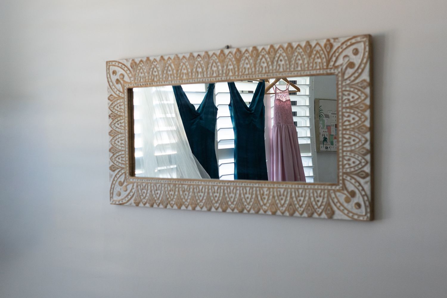 Ornate carved wooden mirror with white distressed finish reflecting formal dresses hanging in a bright room.
