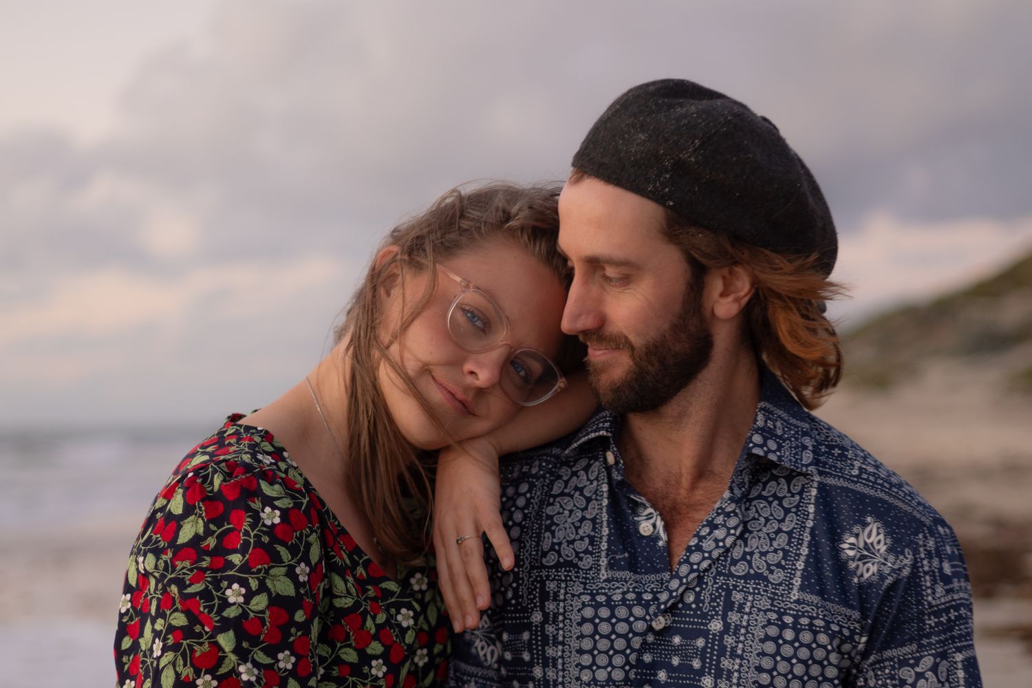 A couple embraces on a beach at sunset wearing floral and patterned clothing in a romantic photoshoot.