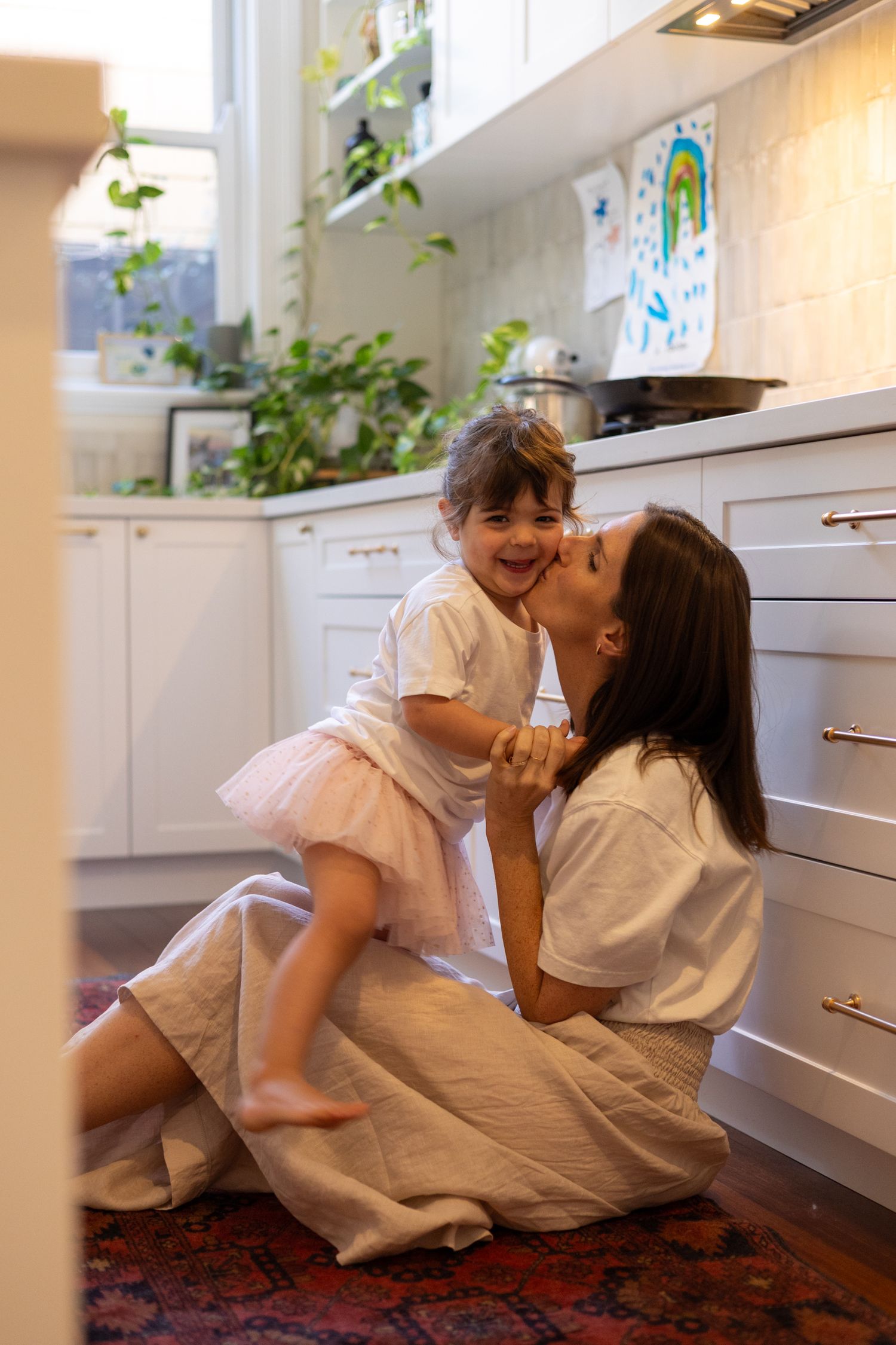 A tender moment shared between parent and child in a bright white kitchen with a red rug and potted plants.
