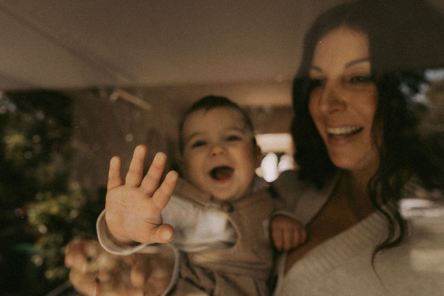 A joyful baby waves at the camera while being held by a smiling woman in a warmly lit indoor setting.