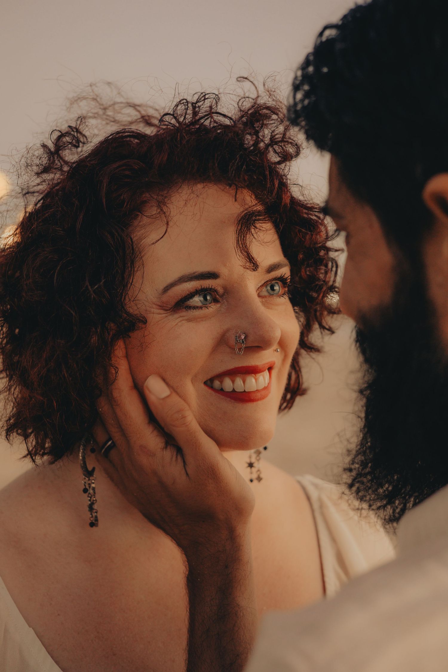 A smiling woman with curly hair and red lips gazes up as a bearded man gently cups her face in a romantic moment.