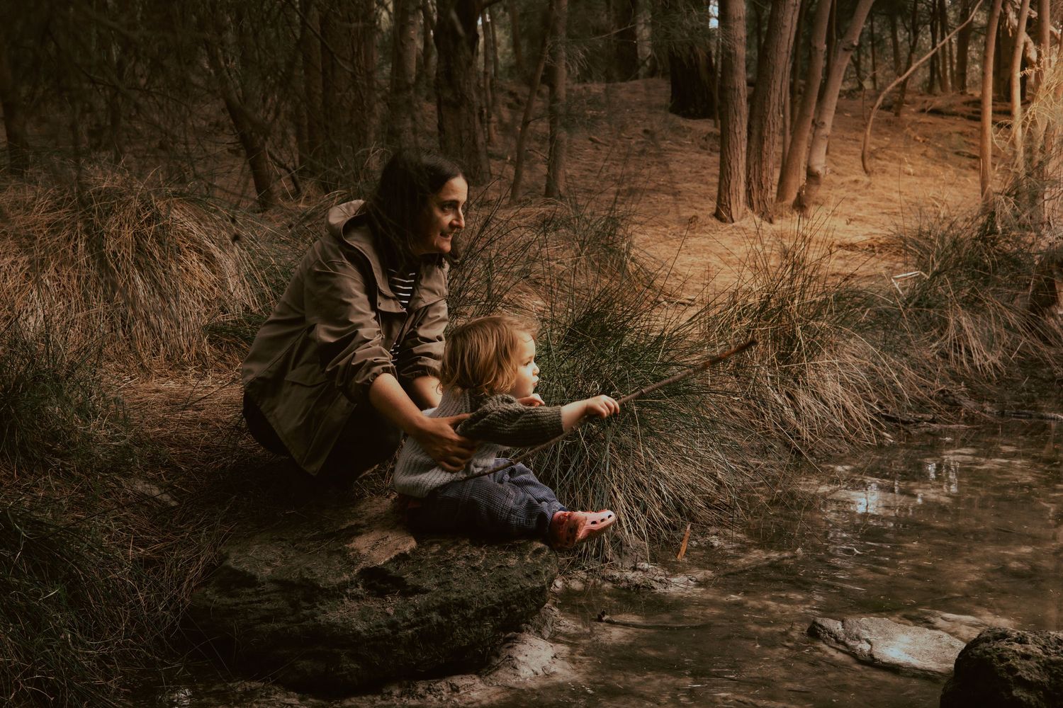 A mother and toddler sit by a forest stream on rocks, exploring nature together in a woodland setting.