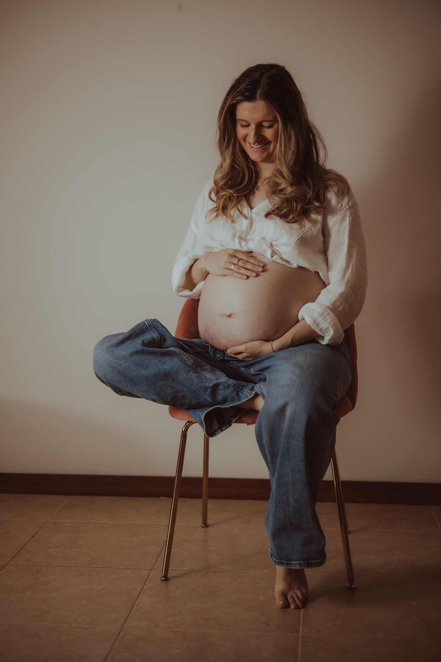 A pregnant person in jeans and white shirt sits on a wooden chair showing their growing belly in a maternity photoshoot series.