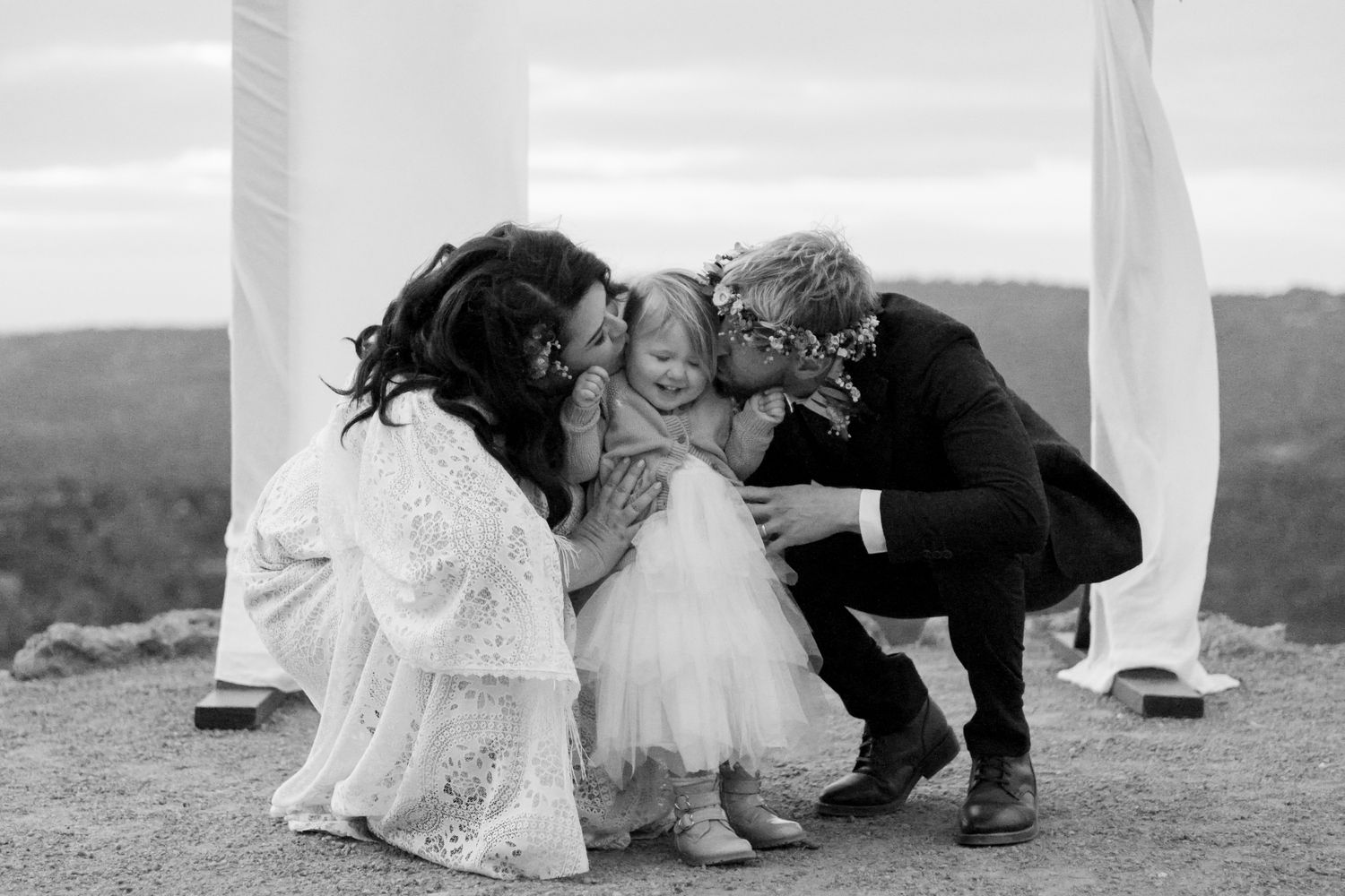 Black and white wedding photo of bride and groom kissing flower girl outdoors at scenic hilltop ceremony.