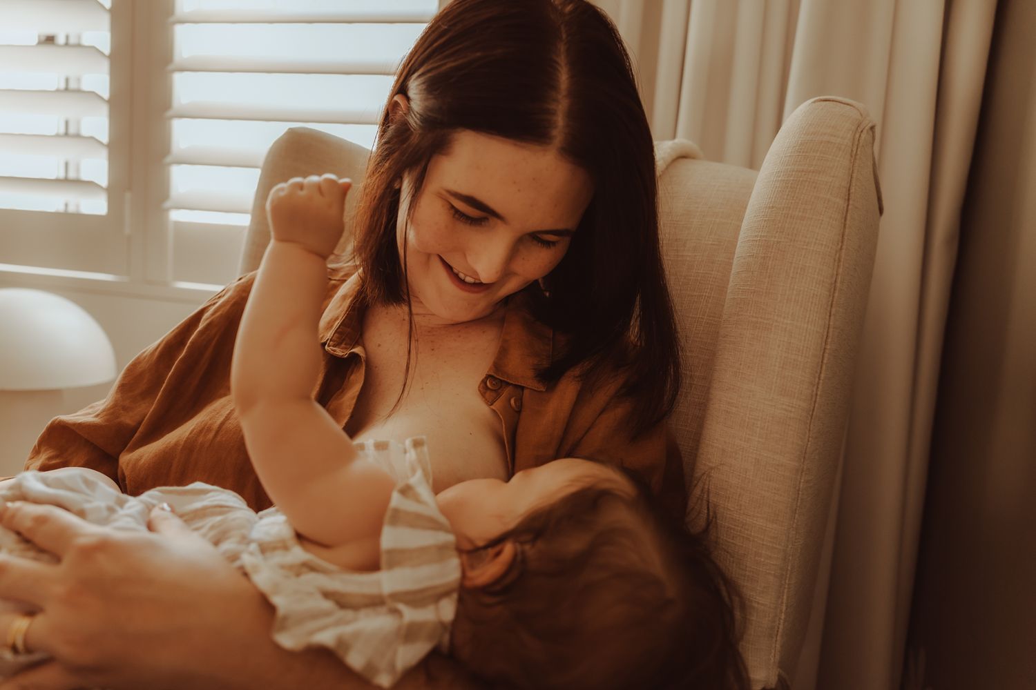 A mother in a white dress tenderly holds and gazes at her newborn baby in a warm, sunlit room.