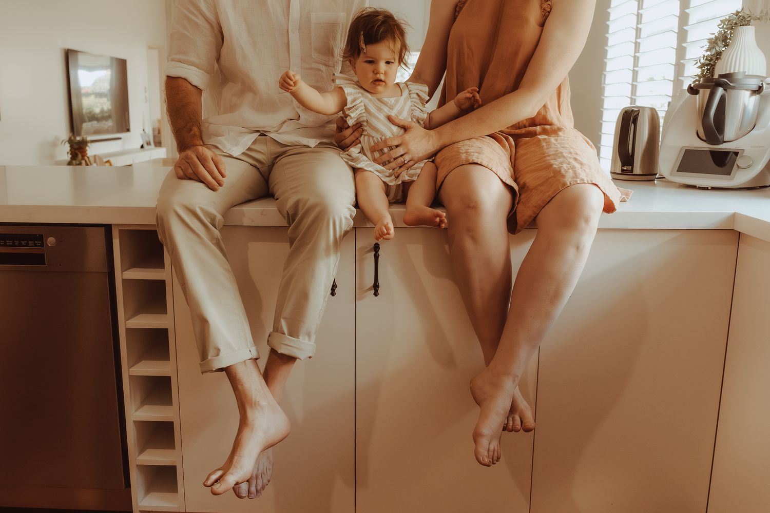 A child sits between two adults on a bathroom counter in warm neutral tones.