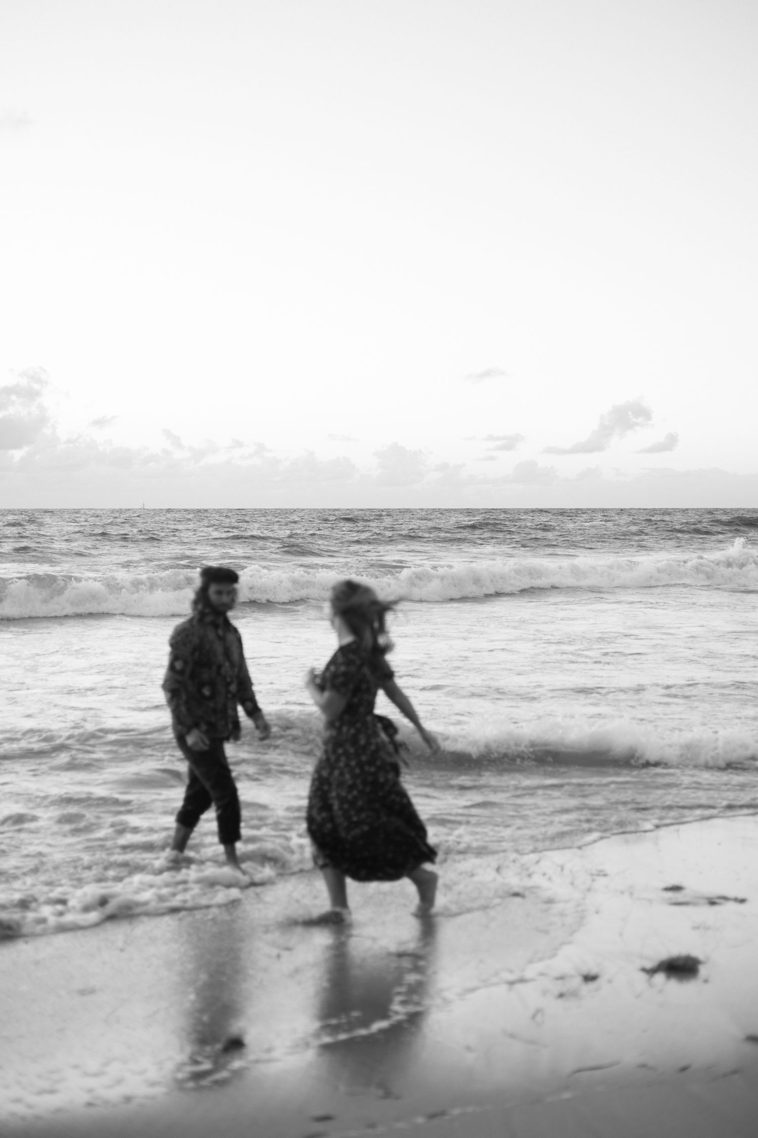 A black and white photo of two people walking along a beach shoreline at sunset. Perth wedding documentary photography, couples, proposal