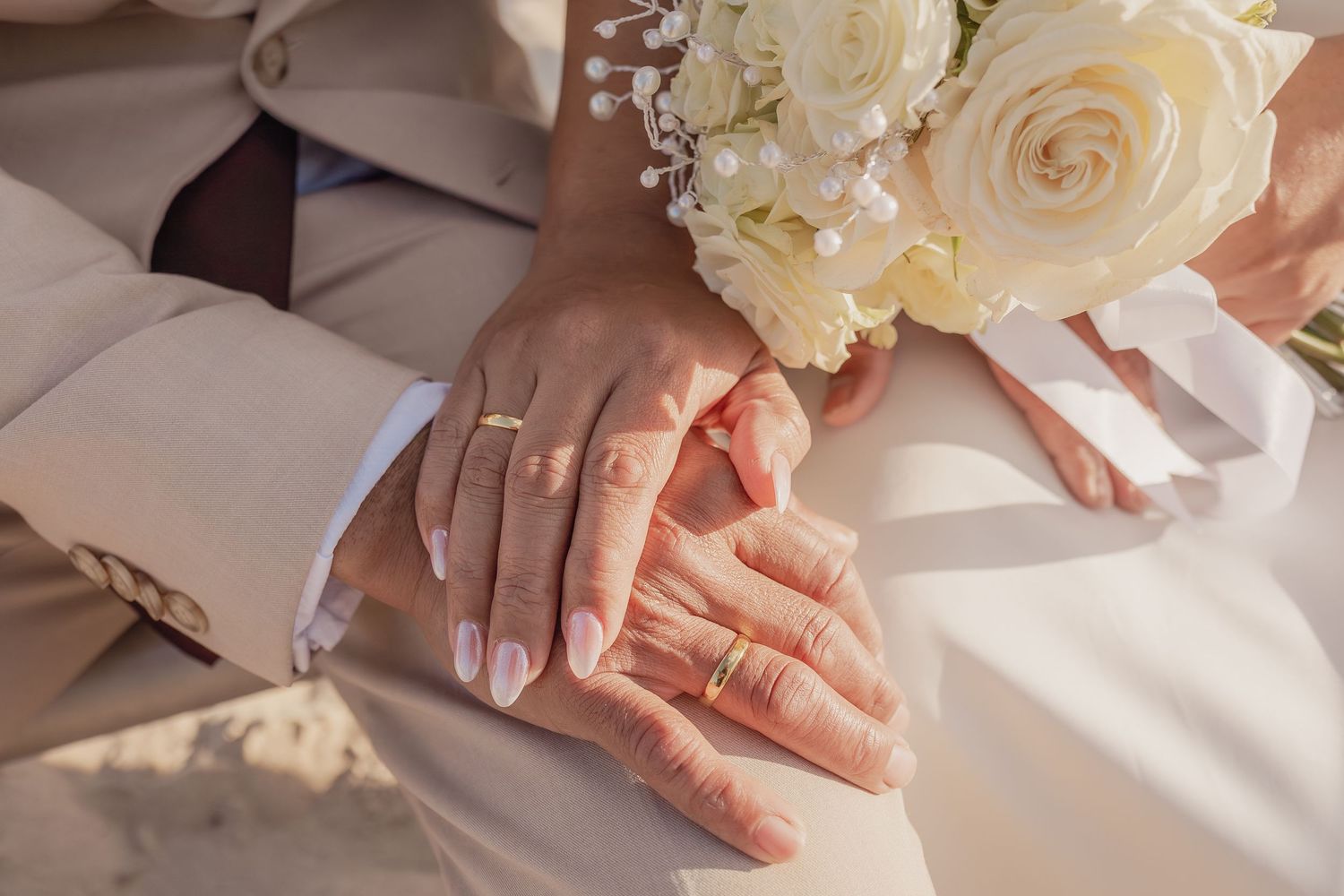 Newlywed couple holding hands with wedding rings and white rose bouquet on wedding day.