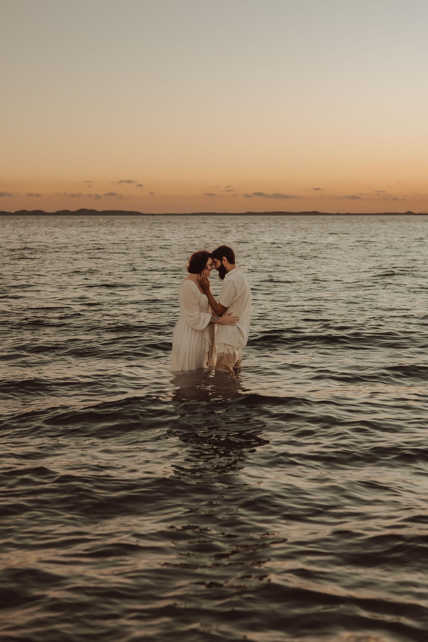 Couple in white attire embracing in ocean water during golden sunset, romantic seaside engagement photo.