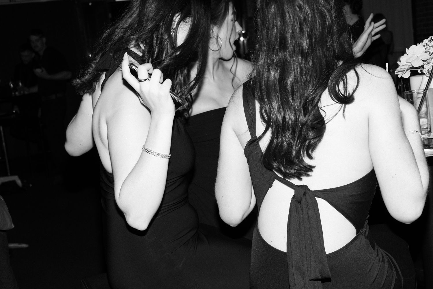 Black and white photo of three people in dark clothing dancing with hands covering their faces at a party.