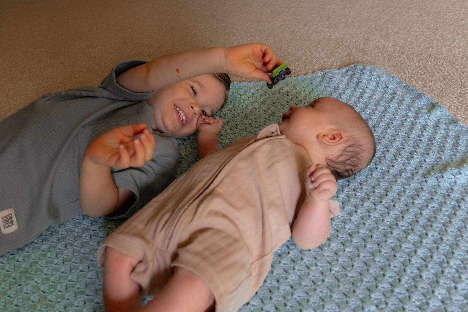 Two babies lying together on a blue textured blanket, one wearing gray and one wearing beige outfits.