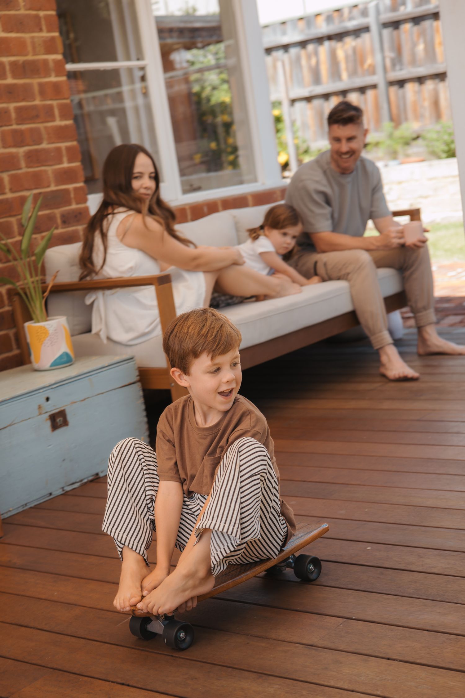 A young boy plays on a wooden deck while adults relax in the background on a sunny day.