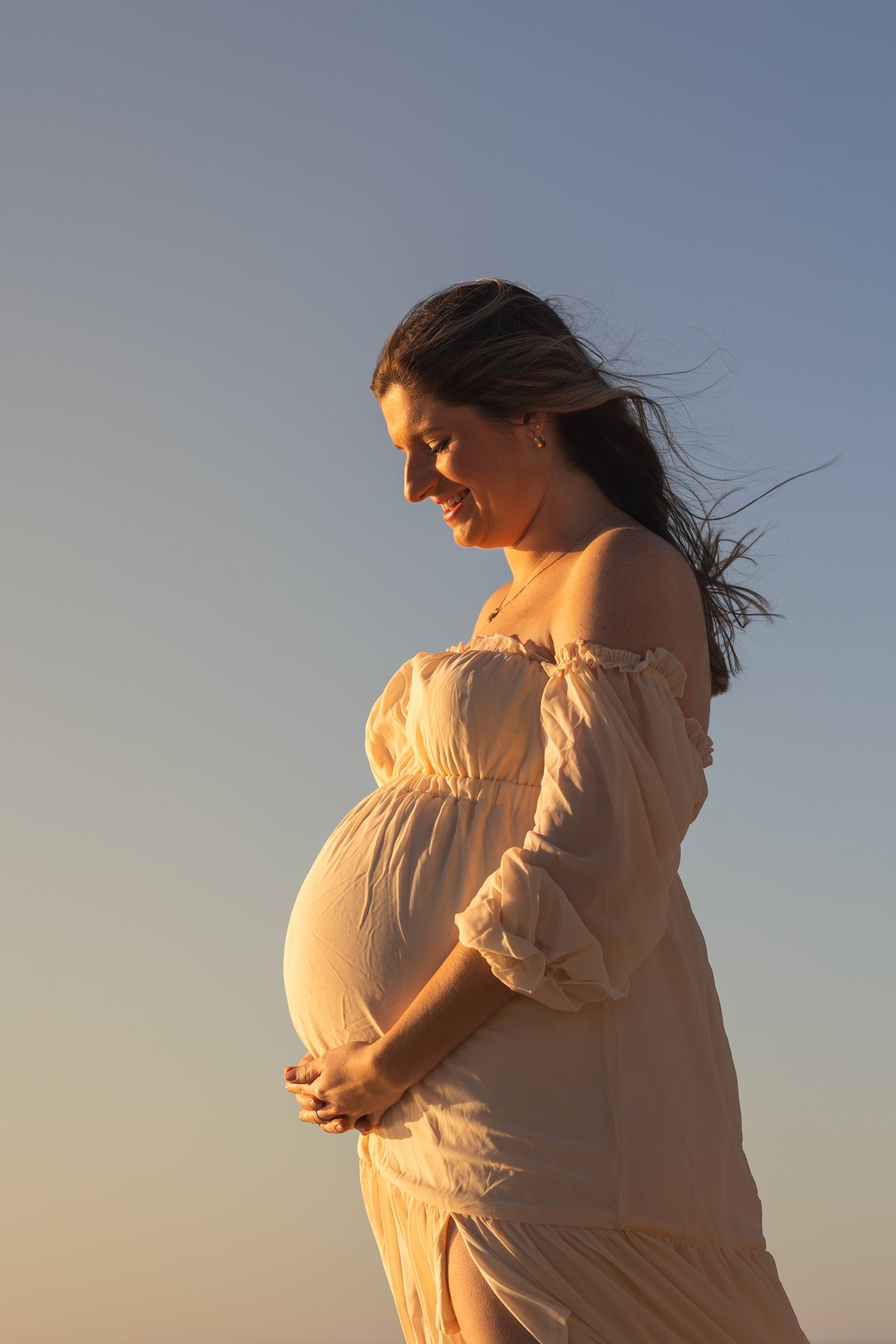 Silhouetted figure in flowing white dress poses against sunset sky in series of artistic maternity photos.