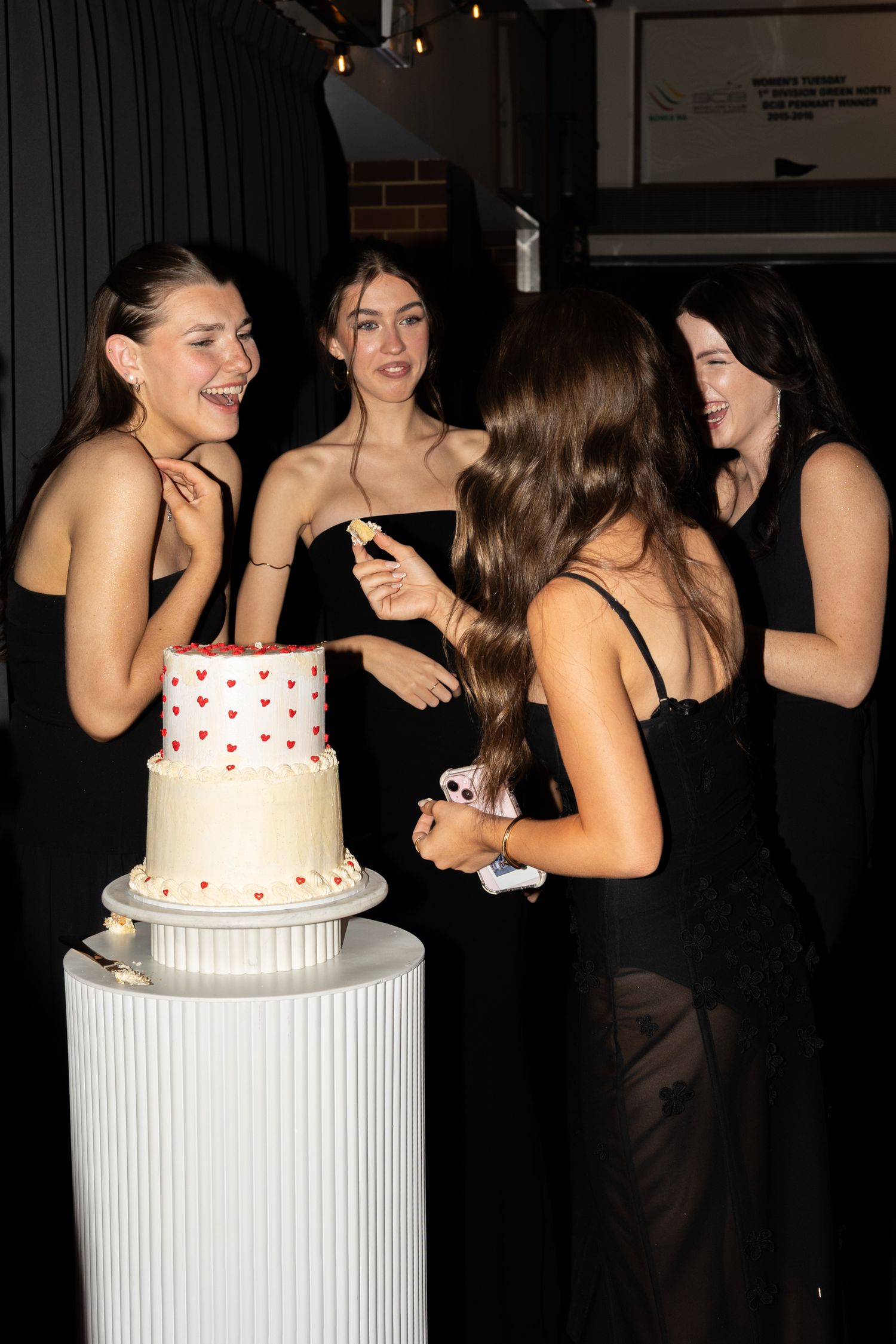 A group gathers around a white tiered cake with red polka dots displayed on a cylindrical pedestal at a celebration.