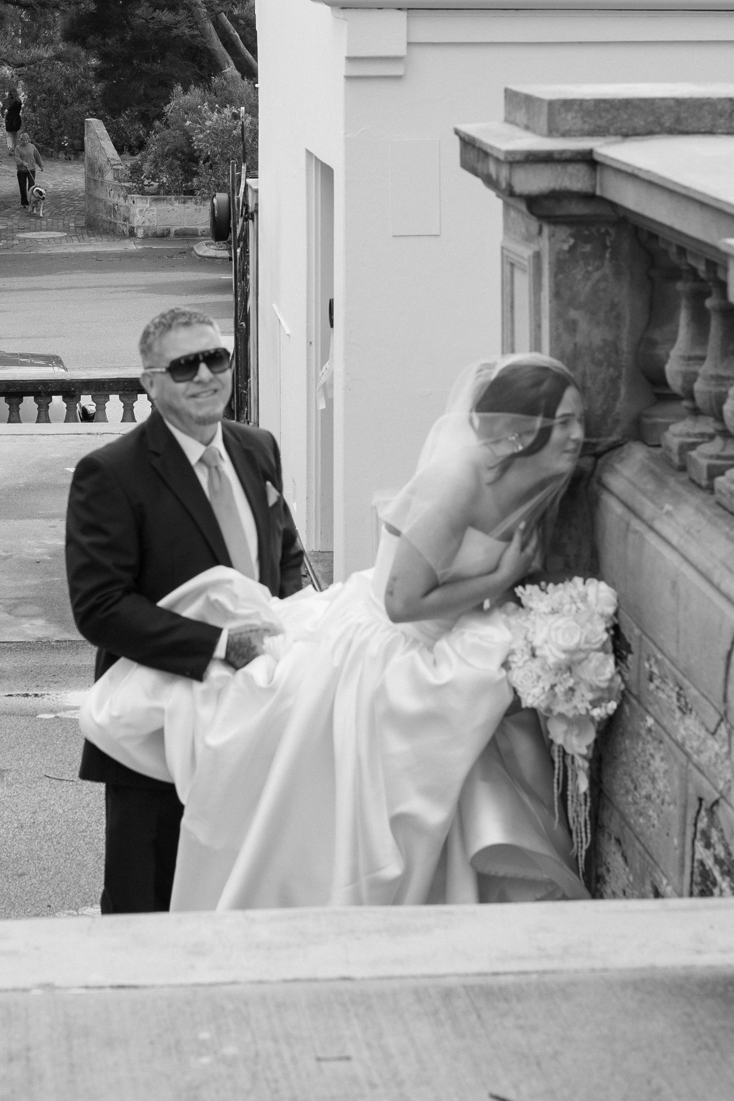 Black and white wedding photo showing a bride in a white dress and veil leaning over a stone balustrade with an attendant nearby.