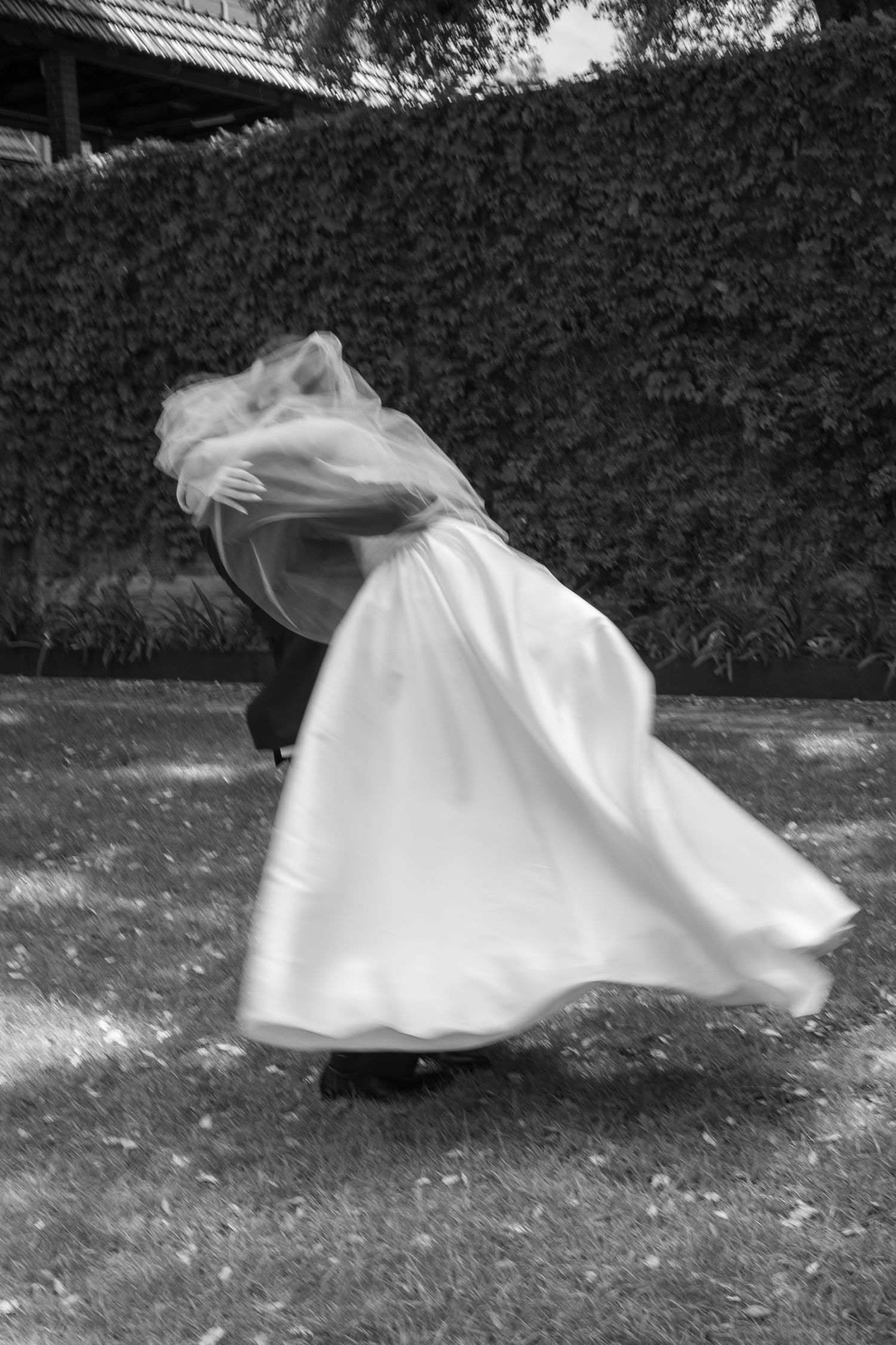 A figure in a flowing white dress leans dramatically in the wind against a garden hedge backdrop in black and white.
