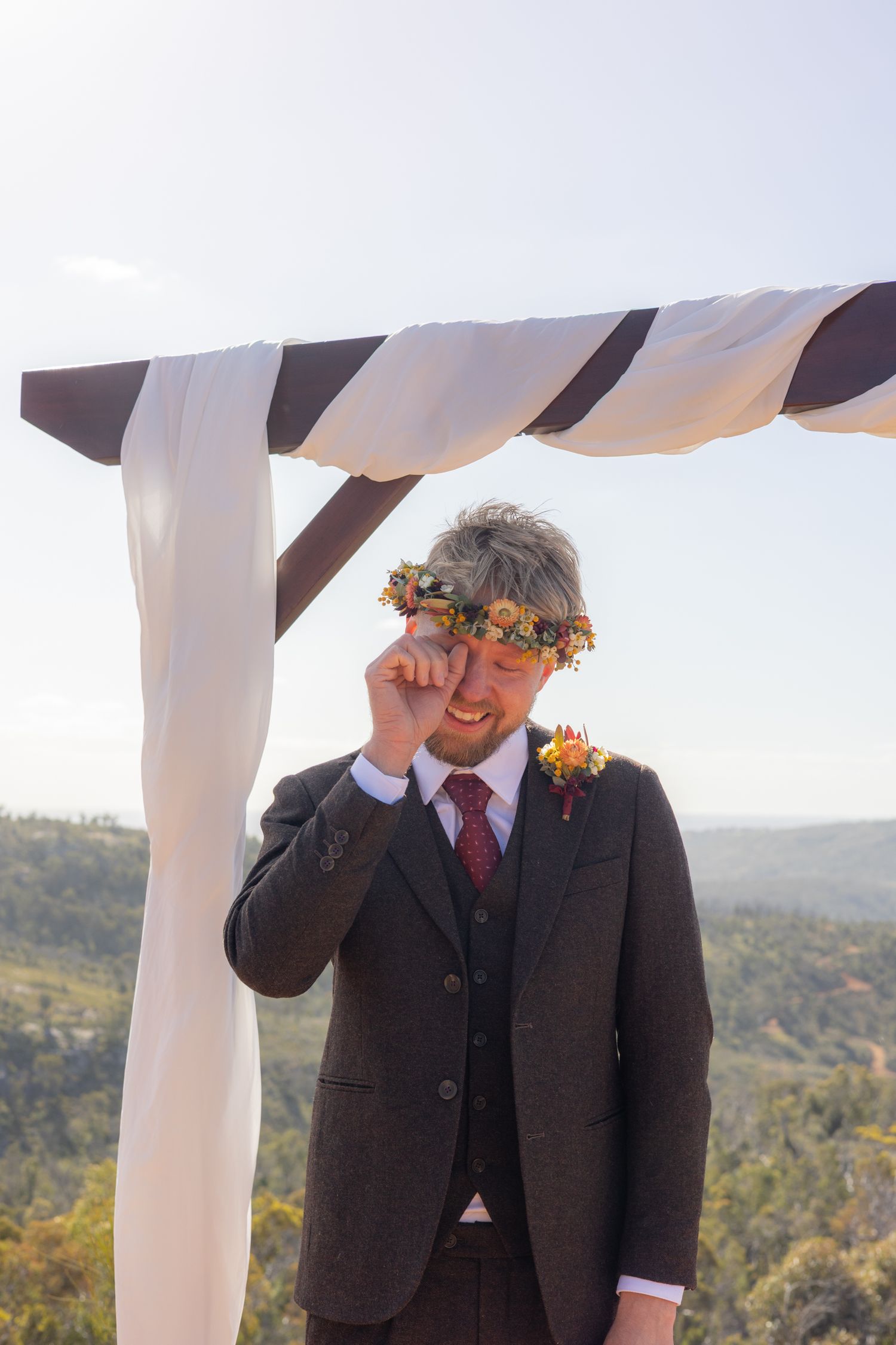 A person in a dark suit and tie with an orange floral boutonniere stands under a white canopy against a scenic hillside view.