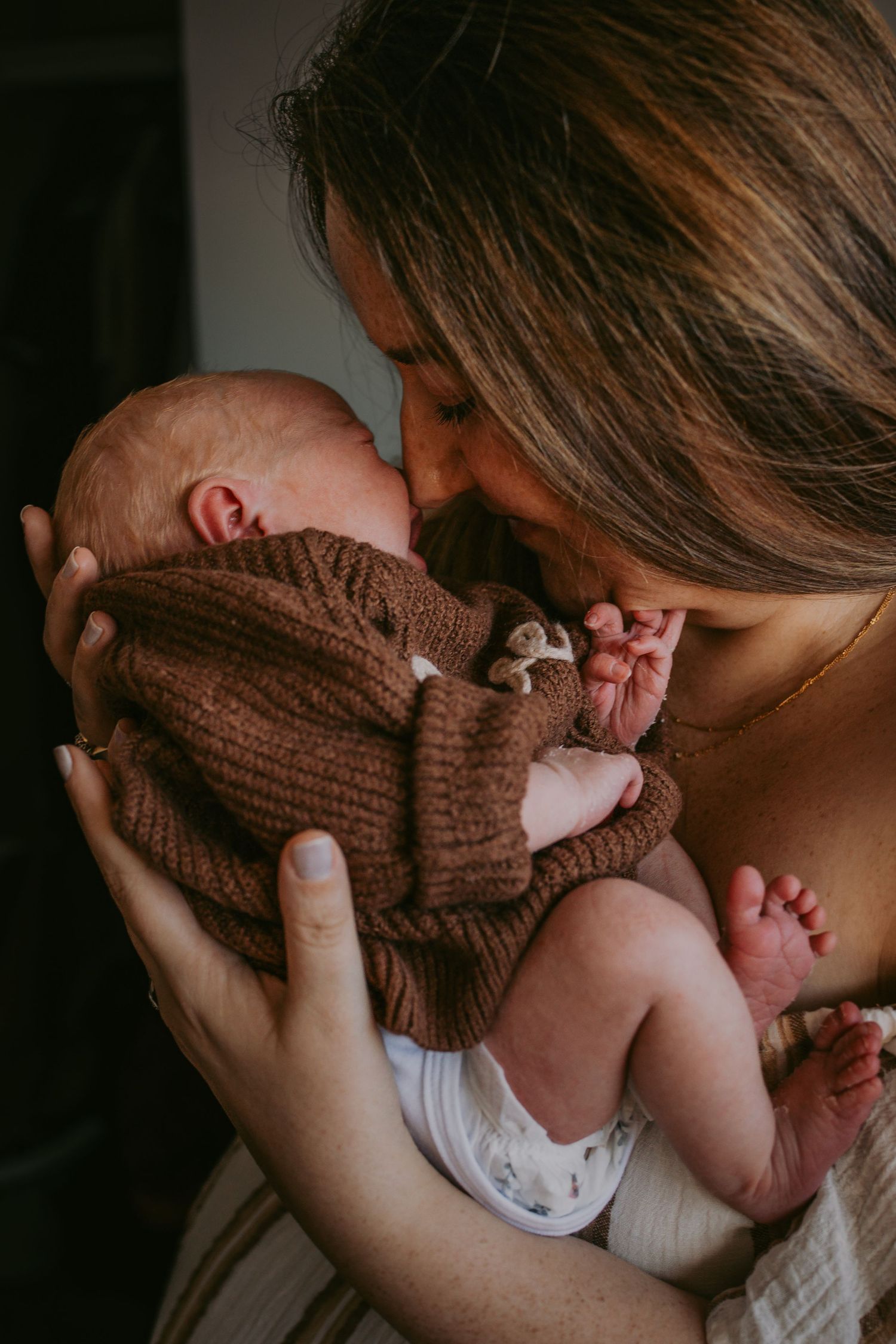 A mother tenderly kisses her newborn baby dressed in a brown knit romper, cradling them gently in her arms.