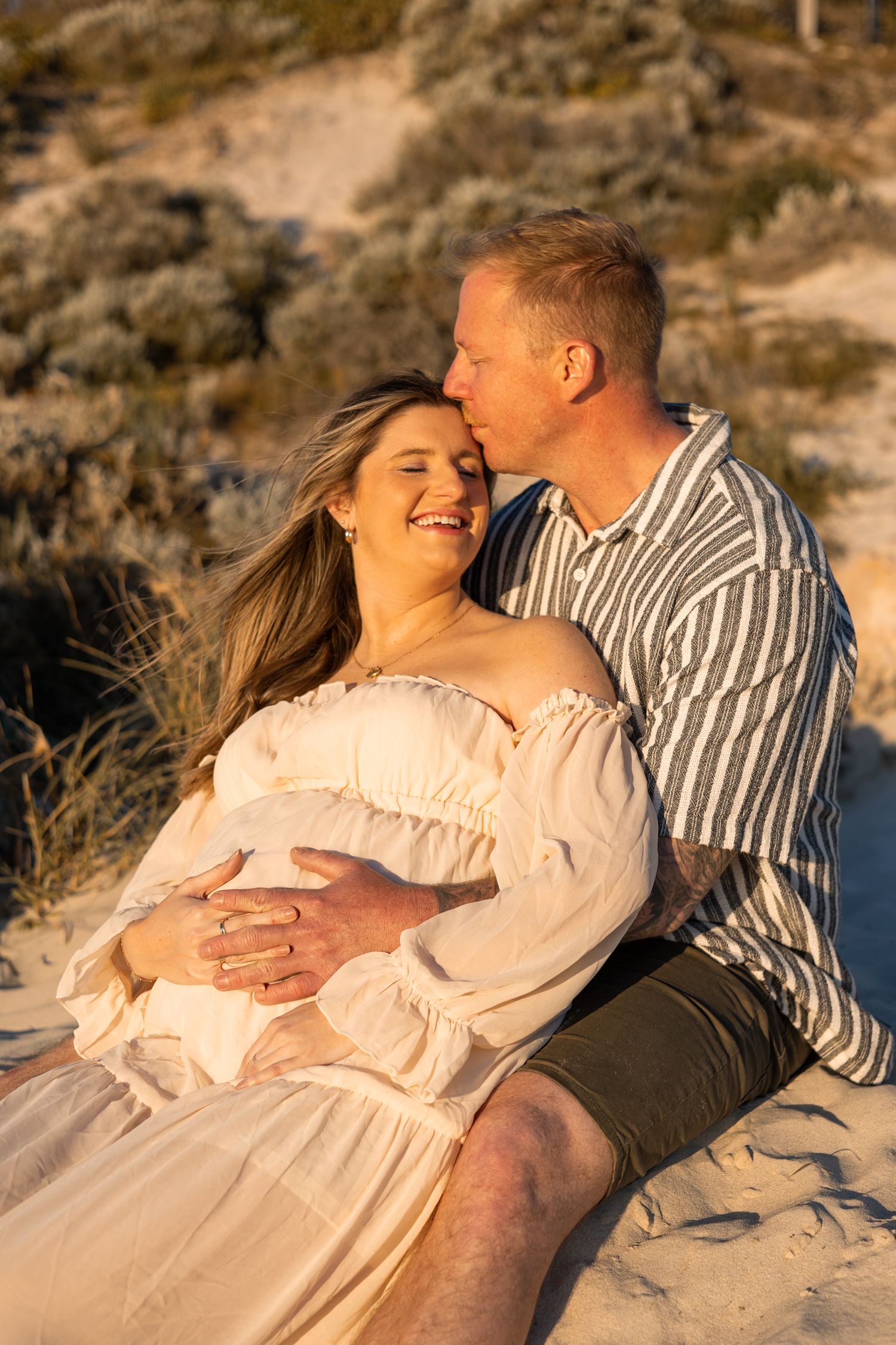 A romantic couple shares tender moments together on sandy dunes at sunset in warm golden light.