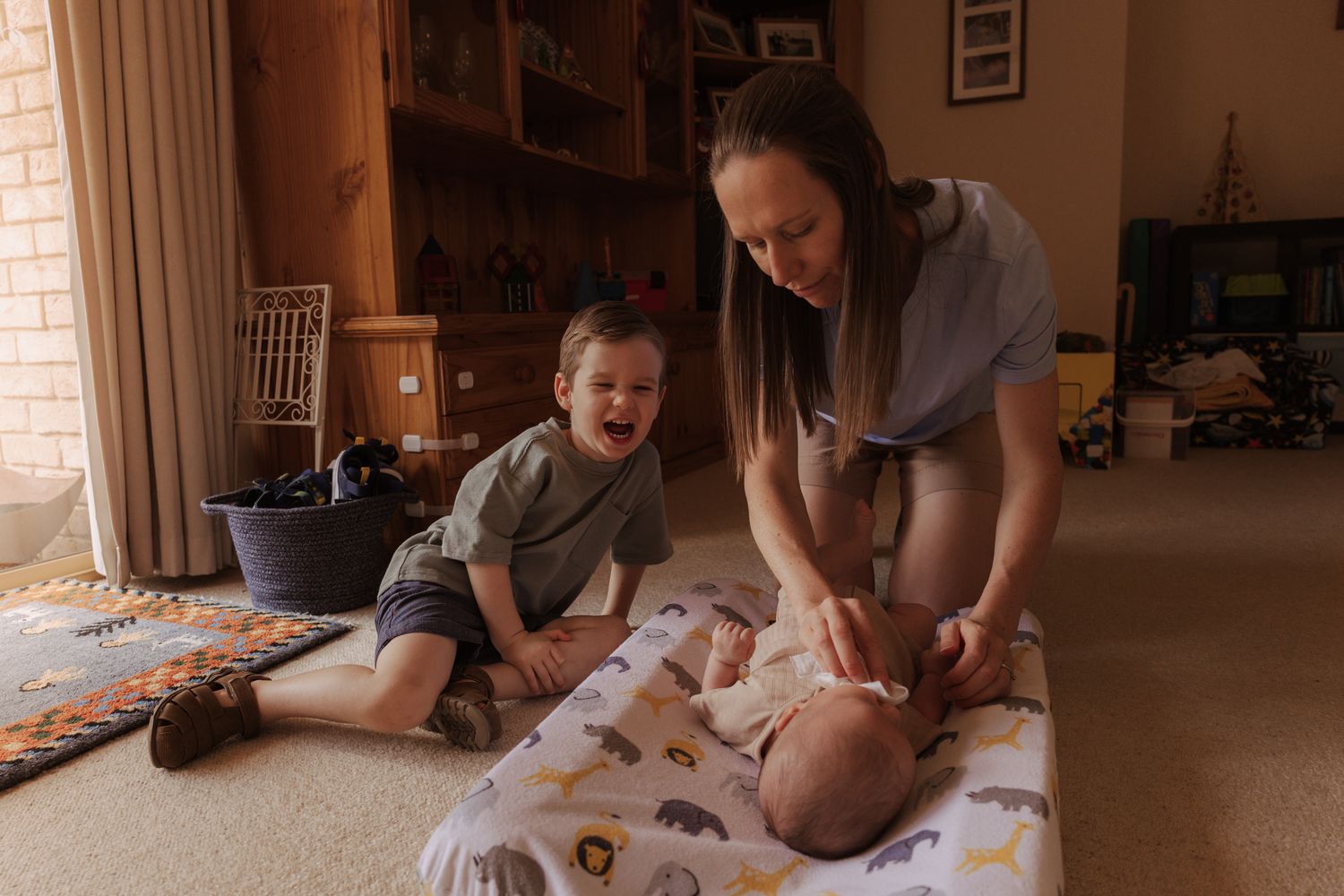 A parent changes a baby's diaper on a blanket while a young child sits nearby on the floor.