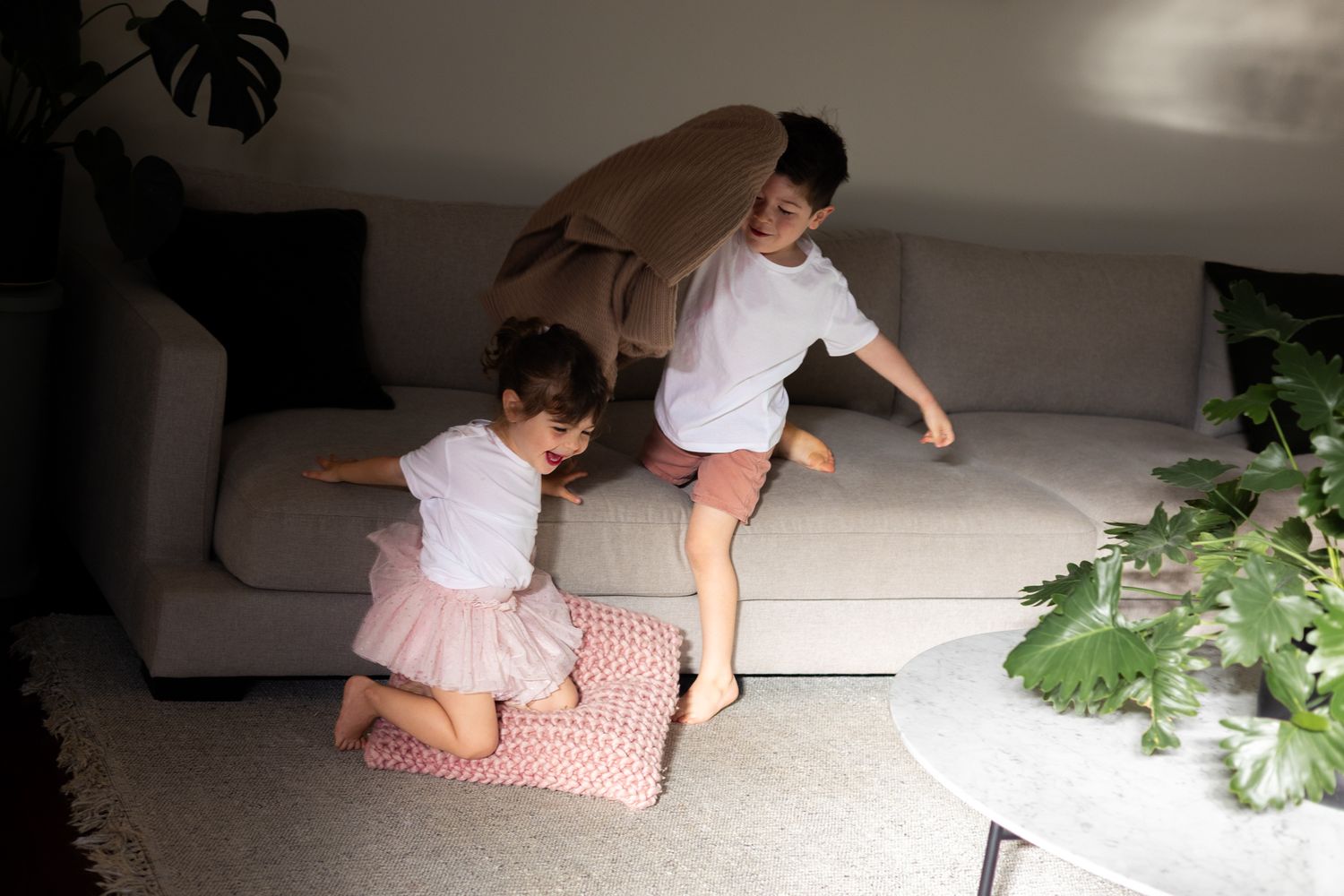 A person in casual attire sits on a white floor near a large potted monstera plant in a bright interior space.