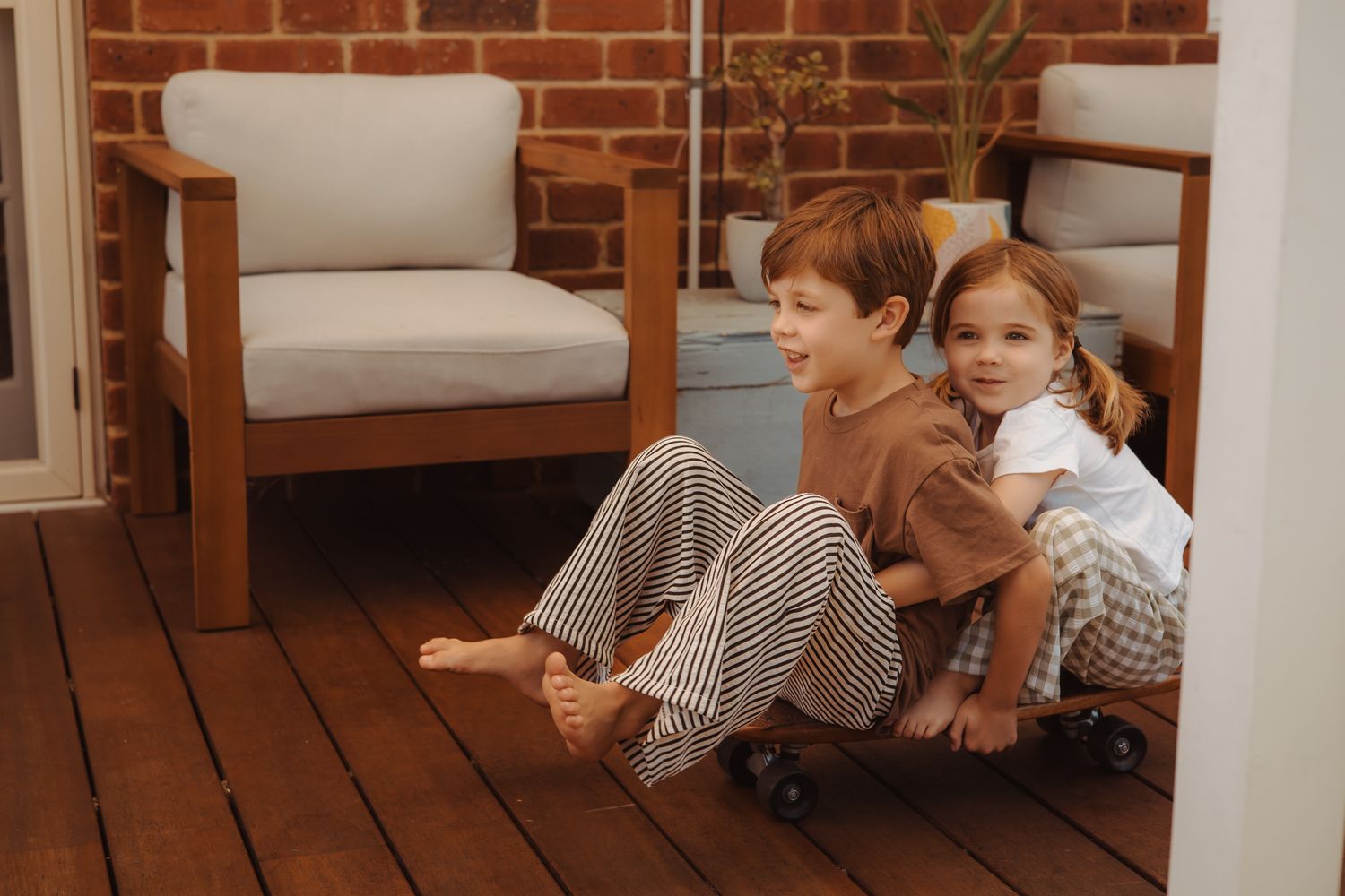 Two children in brown sweaters sit back-to-back on a wooden deck with a cushioned chair in the background.