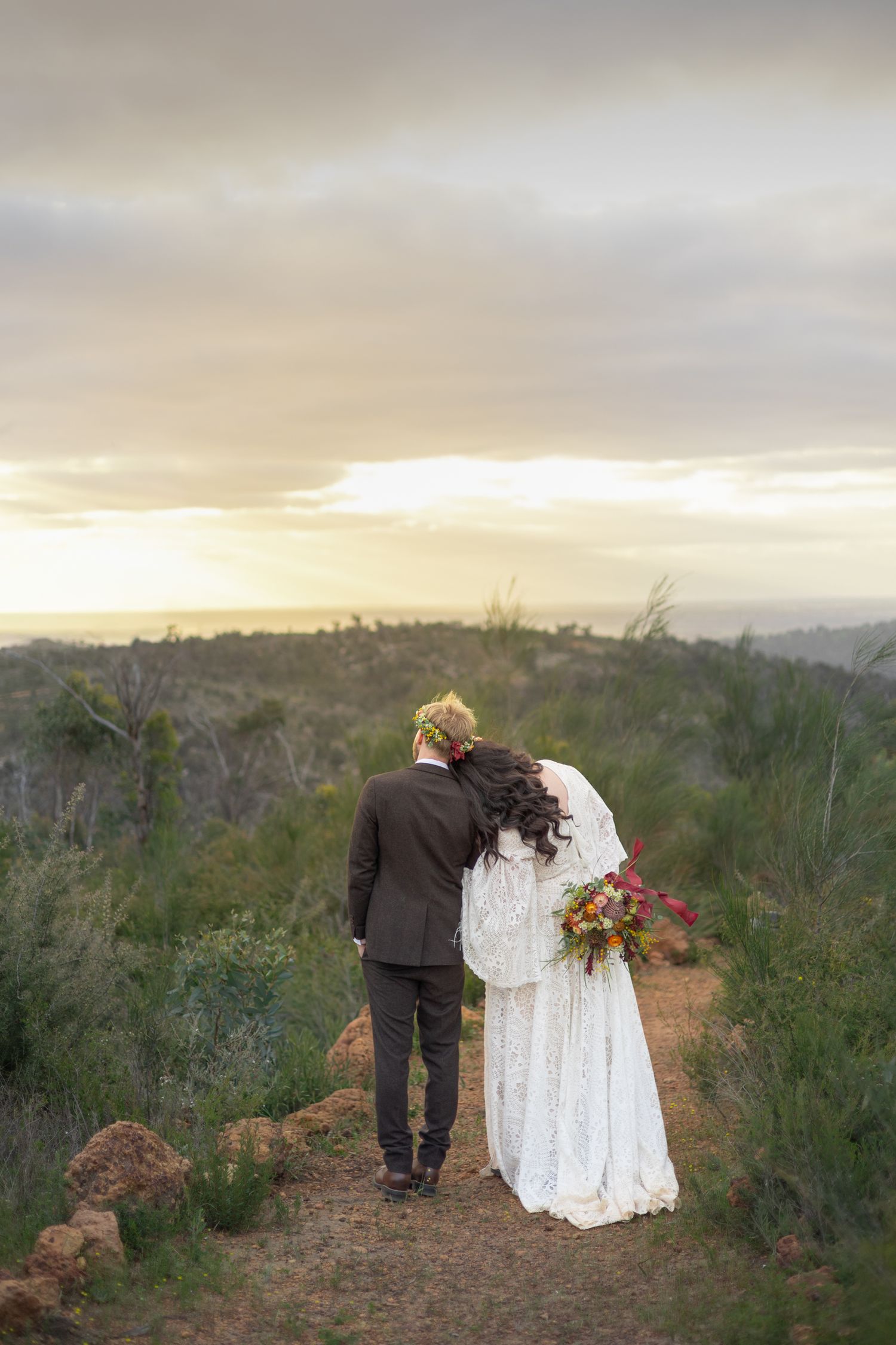 A couple in wedding attire stands on a hilltop path at sunset overlooking a scenic landscape. Perth wedding documentary photography, elopeme