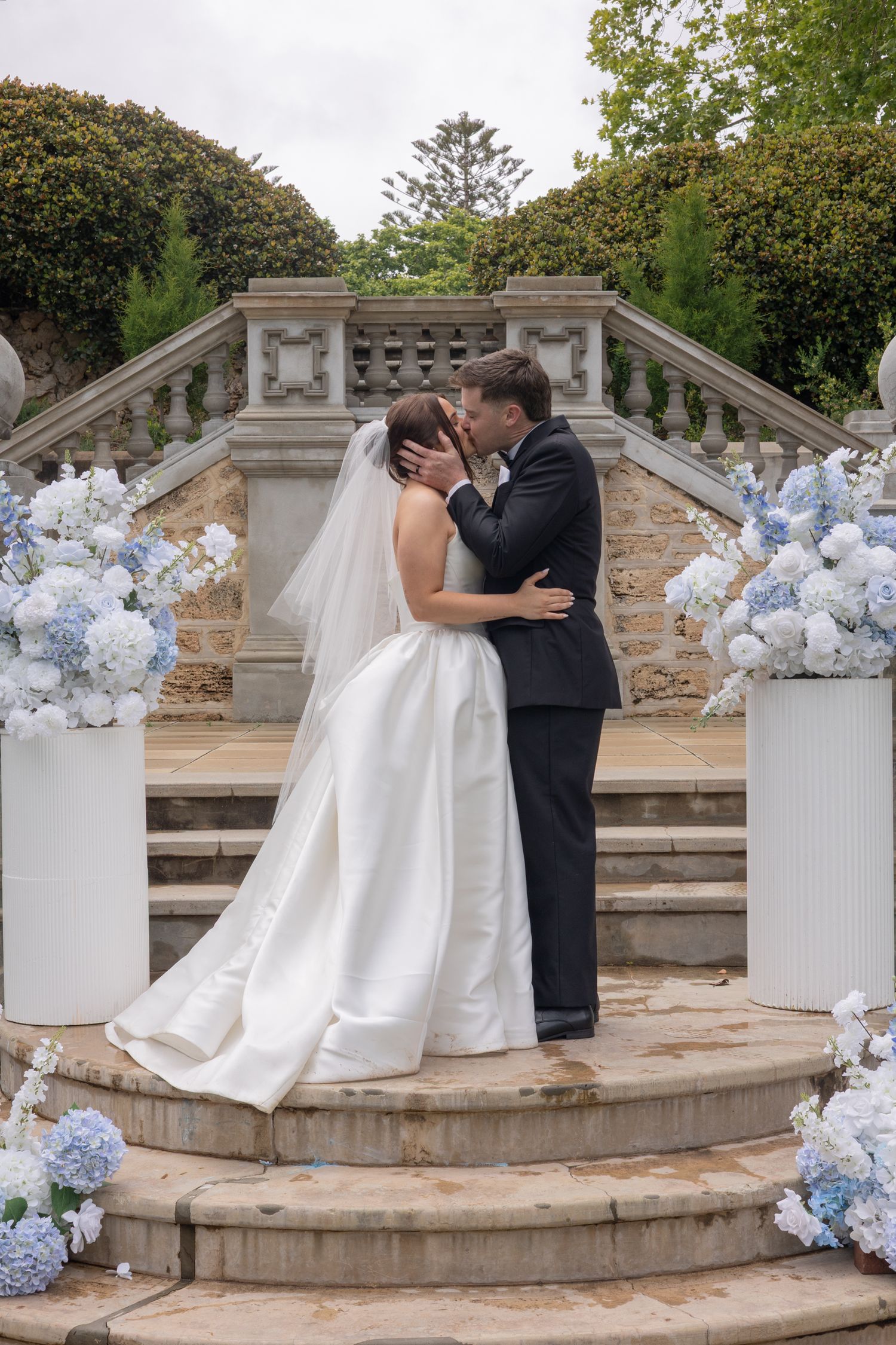 A wedding couple shares a romantic kiss on stone steps decorated with blue and white floral arrangements.