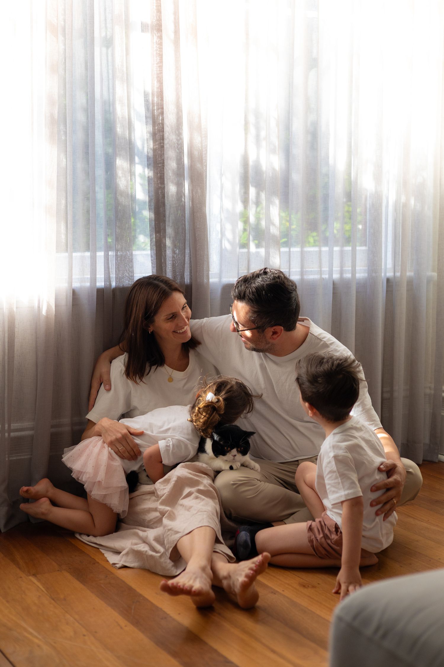 A family shares joyful moments together while sitting on hardwood floor near sheer curtains in a bright room.
