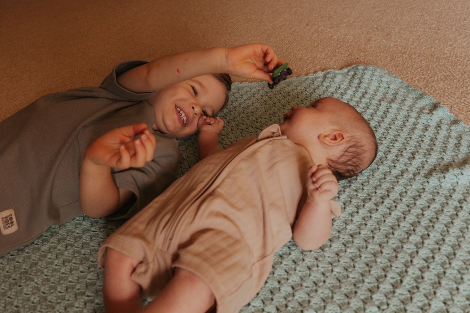 Toddlers lying on a patterned blanket while playing and laughing together.