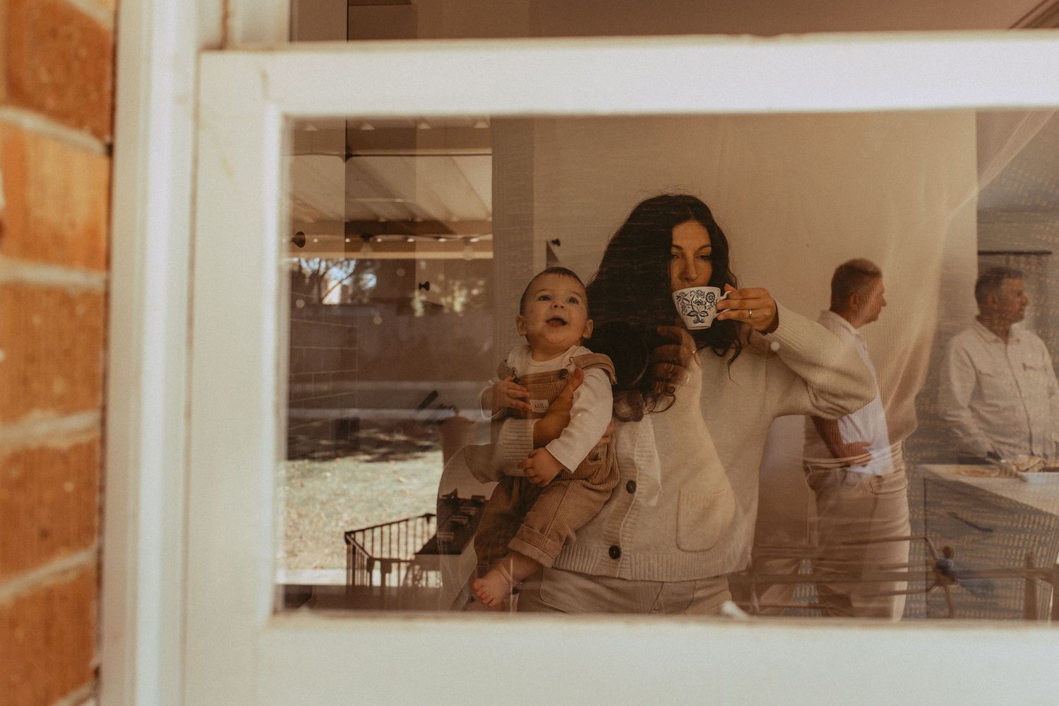 A woman holds a laughing baby while sipping coffee, reflected in a window with a man visible in the background.