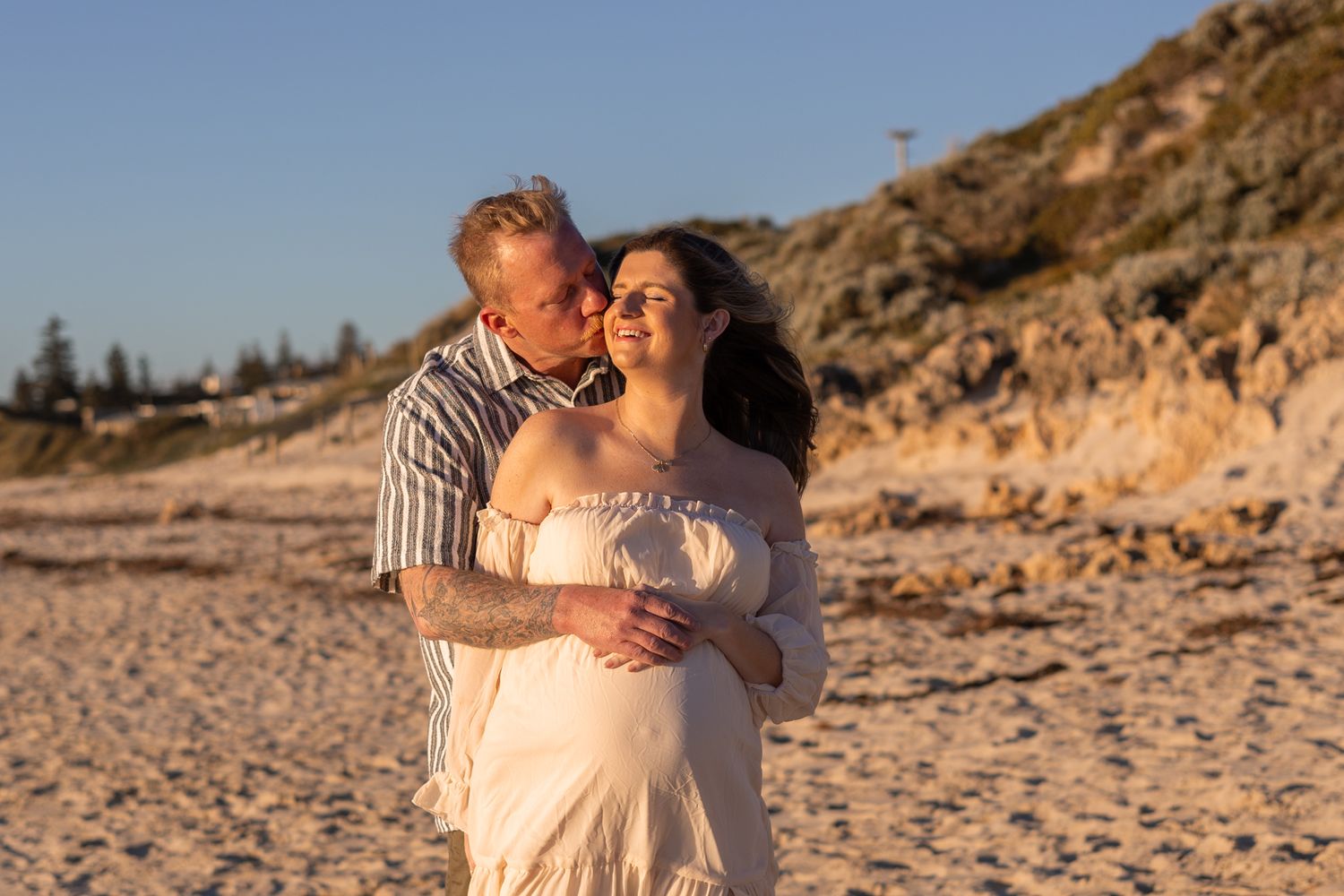 A couple embraces during sunset on a sandy beach with rocky cliffs in the background.