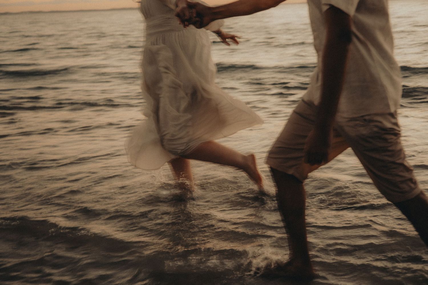 Couple running barefoot through ocean waves at sunset, bride in white dress holding hands with groom in casual attire.