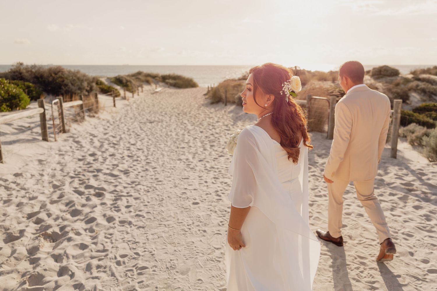 Bride in white dress and groom in beige suit walking on sandy beach path at golden hour sunset.