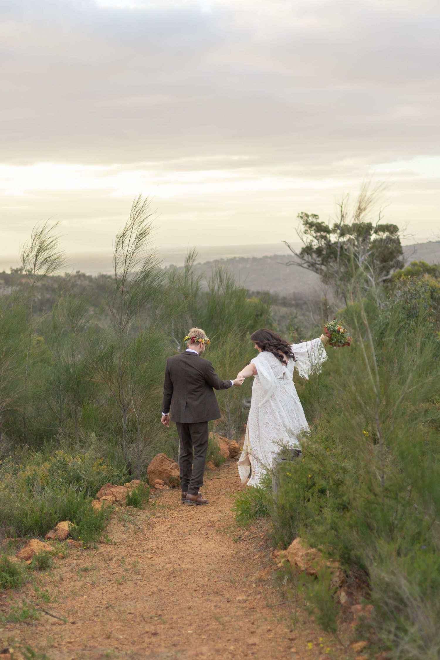 Two figures walk hand in hand down a scenic dirt path surrounded by desert brush at sunset.