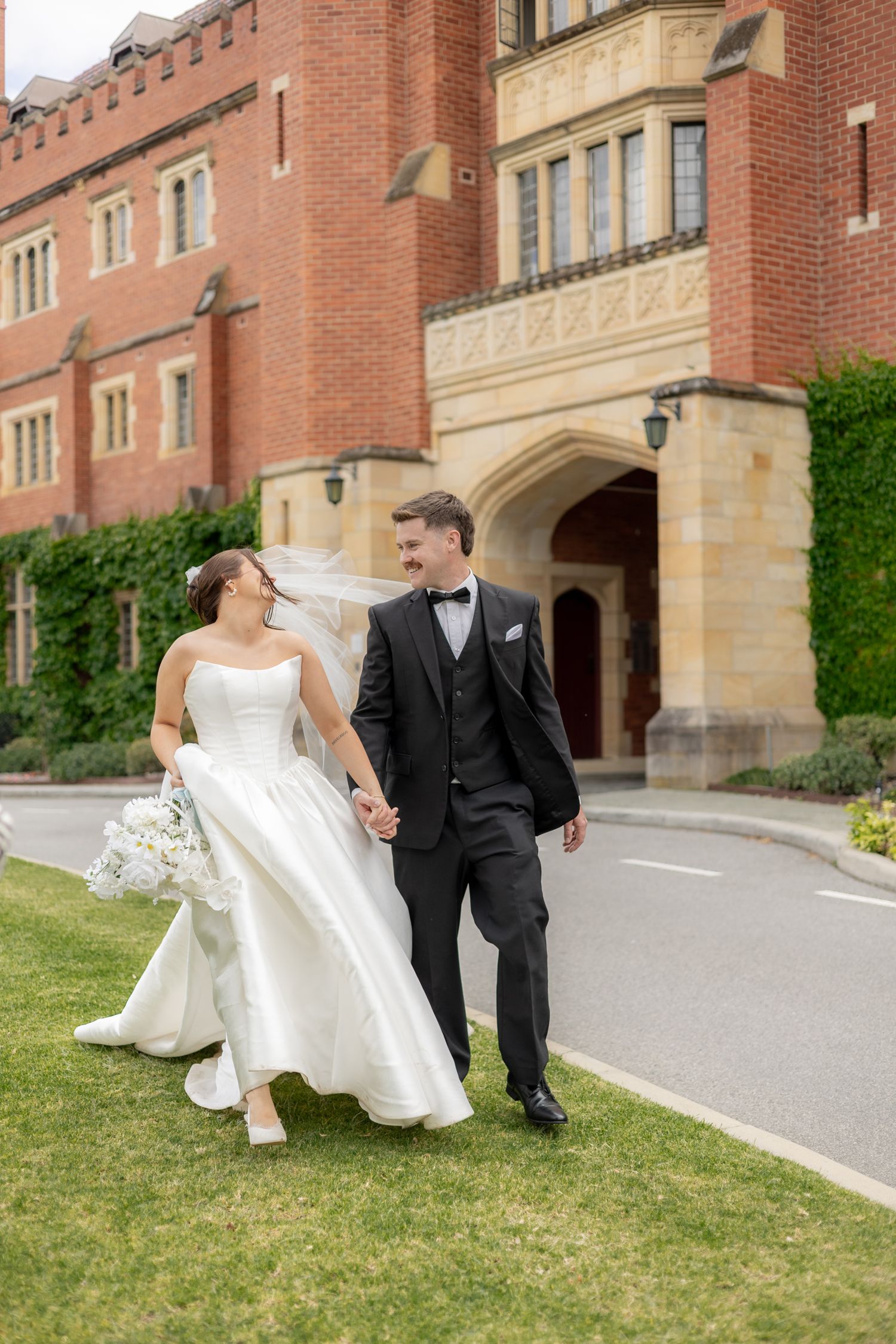 A bride and groom sit together on a grassy hill outside a grand brick university building on their wedding day.