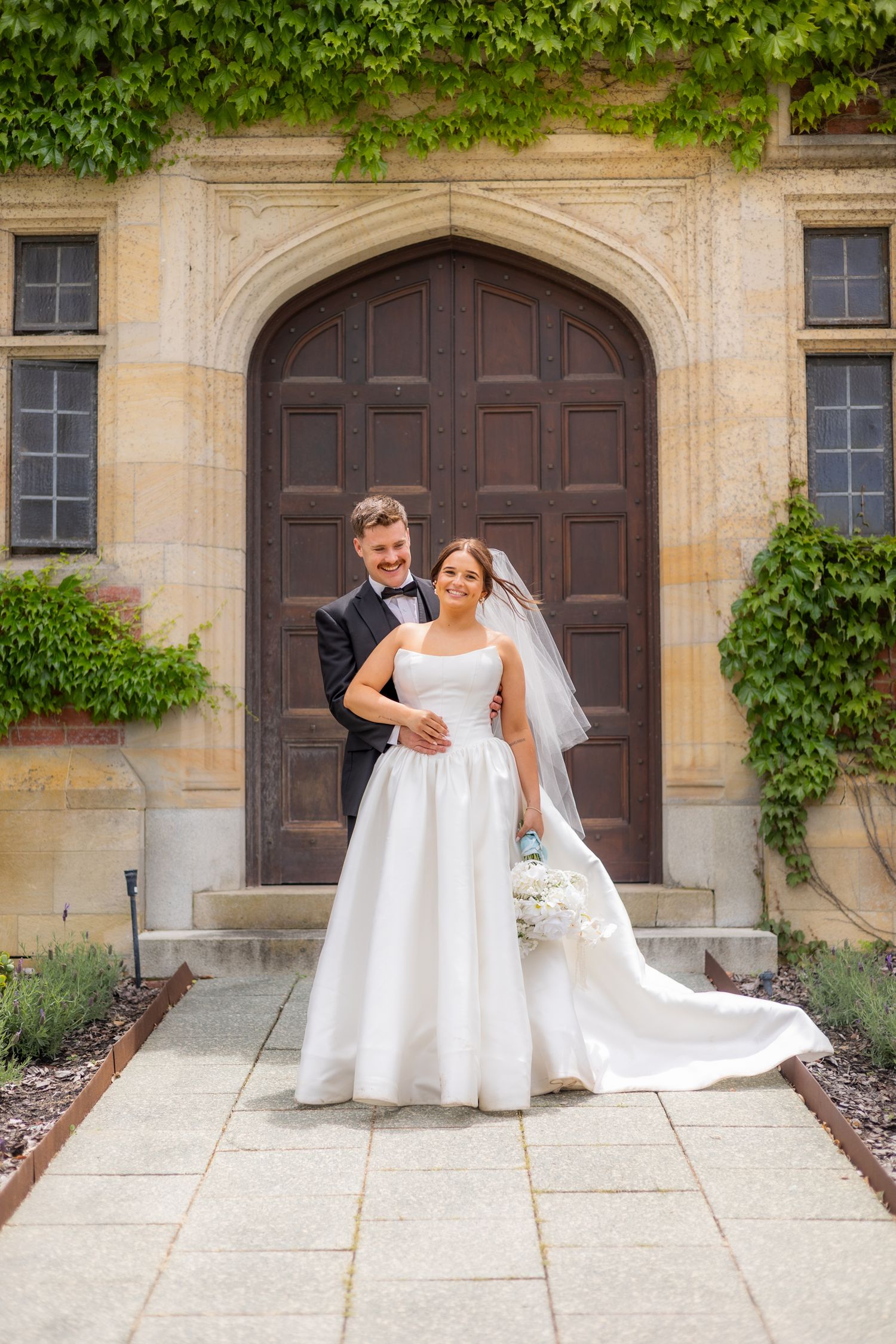 A wedding couple poses playfully in front of an ivy-covered stone building with arched wooden doors. Perth wedding documentary photography